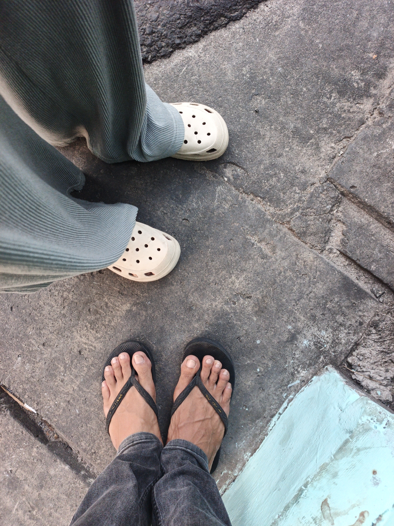 An overhead, medium shot shows two pairs of feet standing on a textured, gray concrete surface. The feet are positioned at different heights within the frame. The upper pair of feet is clad in cream-colored Crocs, worn with loose, teal-green, ribbed pants. Below, the second pair of feet is bare and wears black flip-flops with a simple thong. These feet are covered by dark gray denim jeans. A narrow strip of dark asphalt runs along the top edge of the frame, and a rectangular section of light blue painted concrete is visible in the lower right corner. The scene is consistent with an outdoor urban environment in West Jakarta, Indonesia.