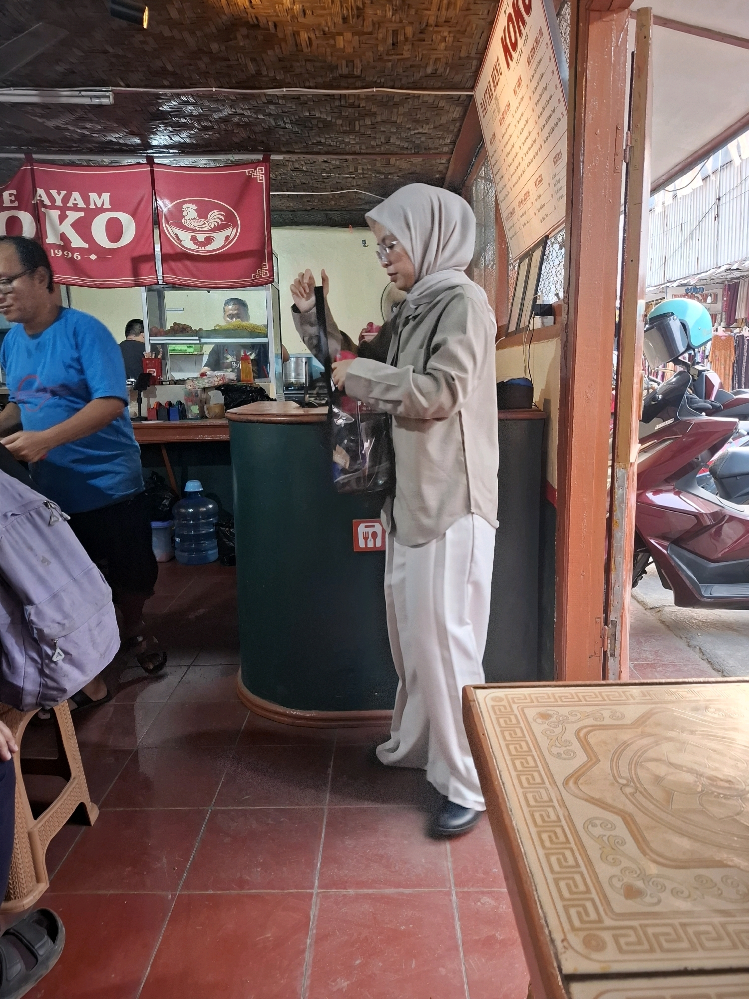 In Cibadak, Indonesia, a woman wearing a beige hijab and light-colored pants stands holding a plastic bag. She appears to be at a food stall or restaurant. To her left, a man in a blue t-shirt and dark pants is partially visible. Behind them, a red banner with the text "AYAM KOKO 1996" and a rooster logo is displayed. A menu is also visible on the wall to the right. In the background, a motorcycle is parked outside. The scene appears to be an outdoor or semi-outdoor setting with a tiled floor.