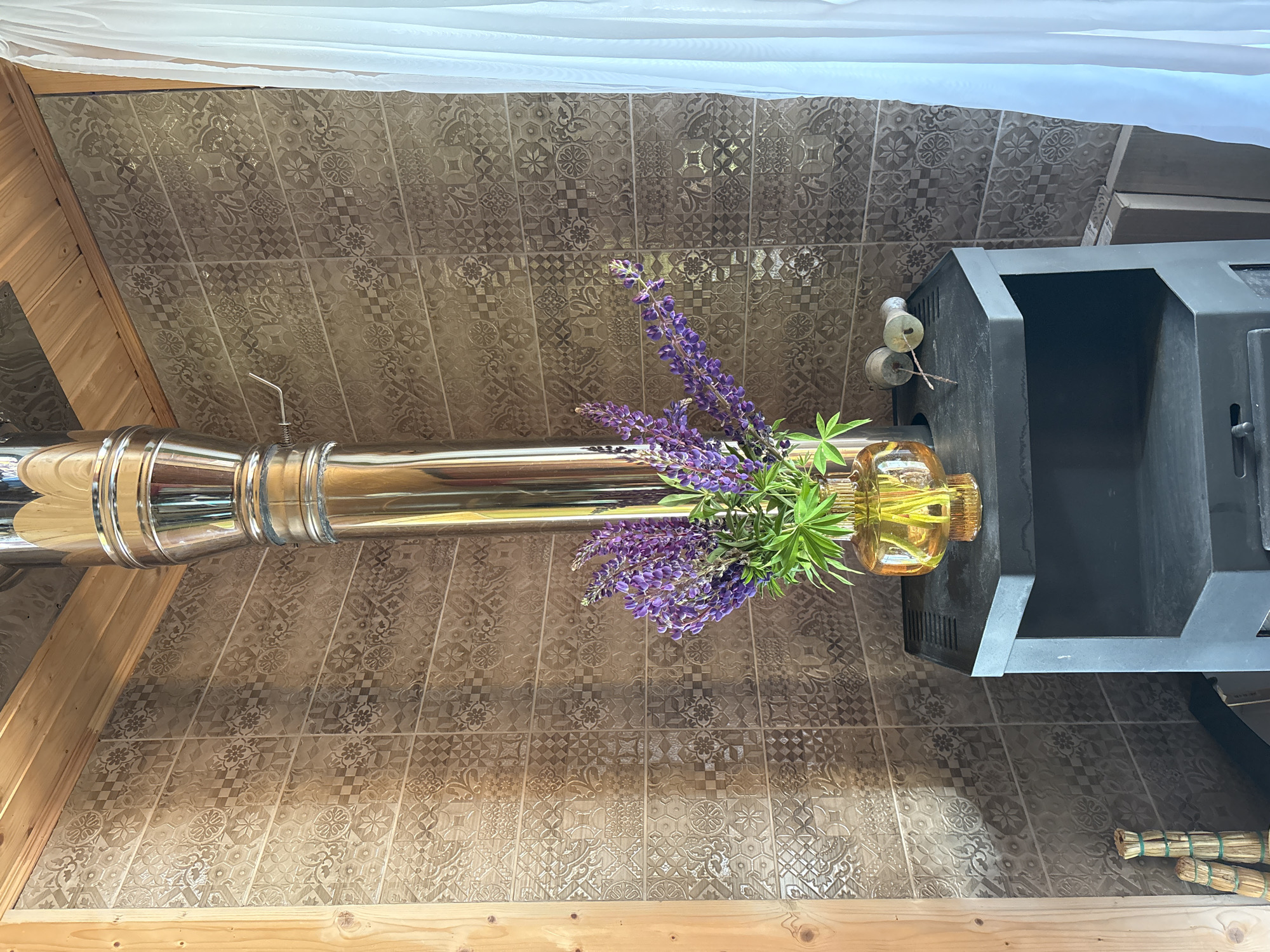 The image shows a corner of a room with rustic decor. A polished metal stovepipe rises vertically from a black stove. The wall behind is covered in brown mosaic tiles with intricate patterns. On top of the stove sits a glass vase filled with purple lupine flowers. To the right of the vase, two small, light-colored objects resembling candle holders are placed. A sheer white curtain hangs on the far right side of the frame. The room appears to be in Lyady, Russia.