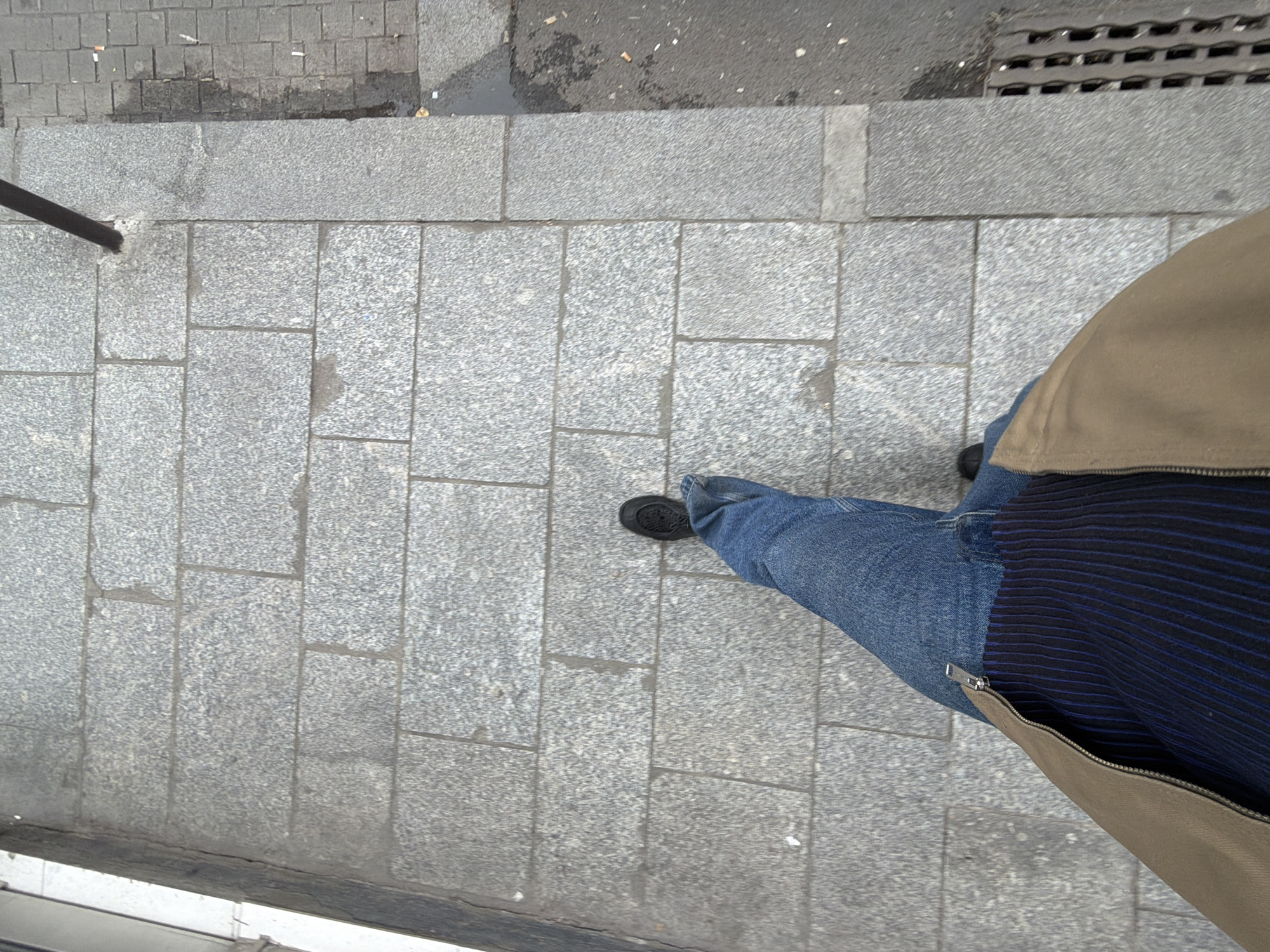 The image is a high-angle shot of a person standing on a granite-tiled sidewalk. The person is wearing blue jeans, black shoes, and a tan-colored jacket with a blue striped shirt underneath. The sidewalk is made of grey granite tiles arranged in a rectangular pattern. Part of a metal pole is visible on the left side of the image, while a metallic grid covers a drain on the upper right side. The area appears to be a street or pedestrian zone in a city, likely in Paris, France, given the granite tiles commonly used in Parisian sidewalks.