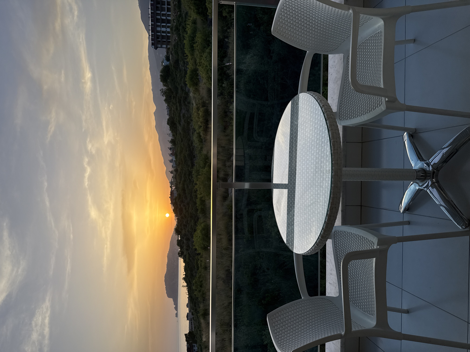 The image shows a sunset over what appears to be a coastal mountain range, as viewed from a balcony. A round table with a white frame and a reflective surface sits in the foreground, surrounded by two white plastic chairs. The balcony features a glass railing. The setting appears to be Alanya, Turkey, suggested by the coastal mountains and building architecture on the left. No people are visible. The only action is implied by the sunset. The environment is warm and serene.