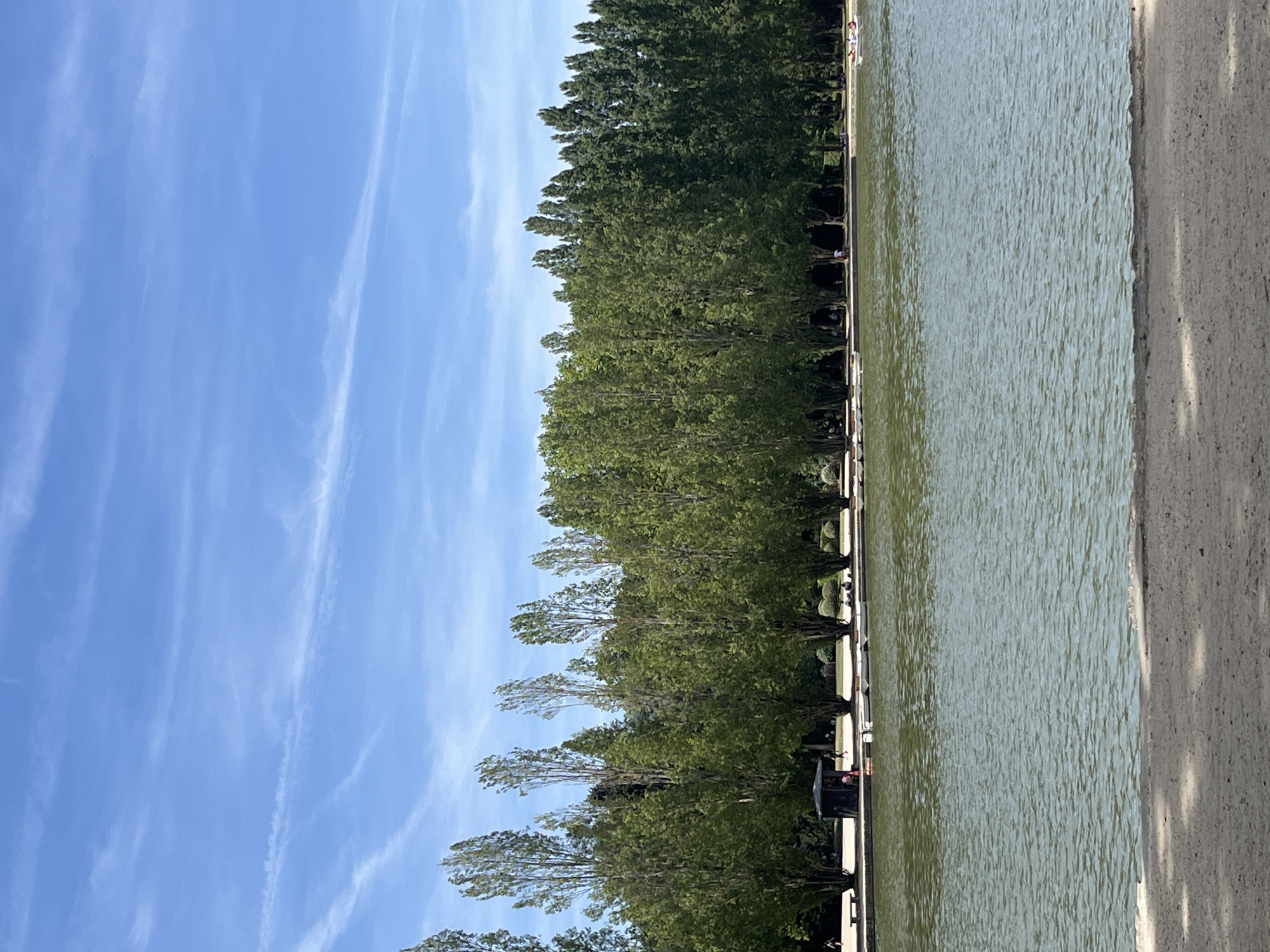 The image shows a wide view of a lake in Antony, France. In the foreground, a sandy embankment leads to the water, which is rippling gently. The middle ground features a line of tall, slender trees along the edge of the lake, with a small brown structure visible amongst them. Several small boats are moored near the shore, and a few people can be seen walking or sitting near the trees. In the background, a dense line of trees forms a backdrop to the scene, and a bright blue sky with scattered white clouds stretches above. The overall impression is one of a peaceful park setting on a clear day.