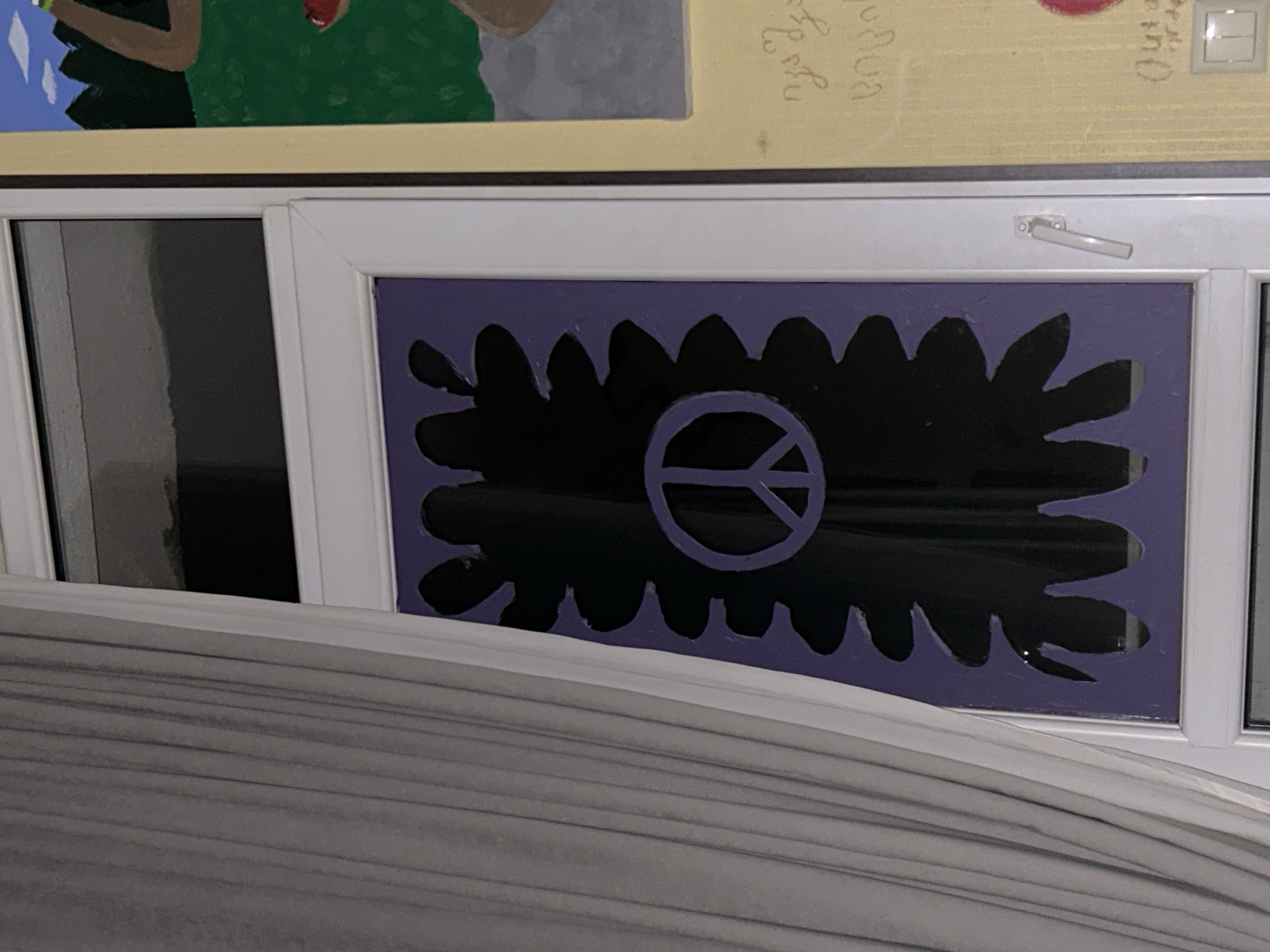 A peace sign is displayed on a window. The peace sign is purple and black, set against a purple background that is cut into a scalloped edge. The window is framed in white. To the left of the window, a grey curtain is partially drawn. To the right of the window, a yellow wall is visible, with some colorful artwork. The artwork appears to be a painting or mural with green and brown figures. There are also some handwritten words or symbols on the yellow wall in what appears to be a foreign language. The image seems to be taken indoors, possibly in a home or art studio.