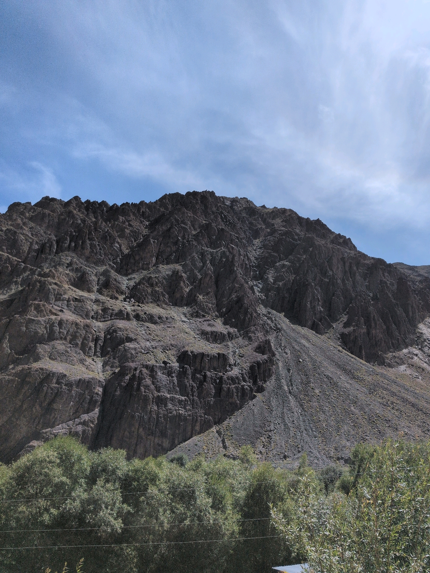 The image shows a mountainous landscape with a rocky, barren slope dominating the upper half. The mountain is dark gray and textured, with visible strata and erosion patterns. The sky above is a bright blue with thin, wispy white clouds. In the foreground, there is a band of lush green vegetation, appearing as trees and bushes, suggesting a valley or lower elevation. The foliage is dense and textured, with sunlight filtering through the leaves. There are also thin, horizontal lines visible in the lower portion of the image, possibly wires. The scene appears to be a natural landscape, likely in a dry, mountainous region, with a contrast between the arid mountains and the greener vegetation at their base.
