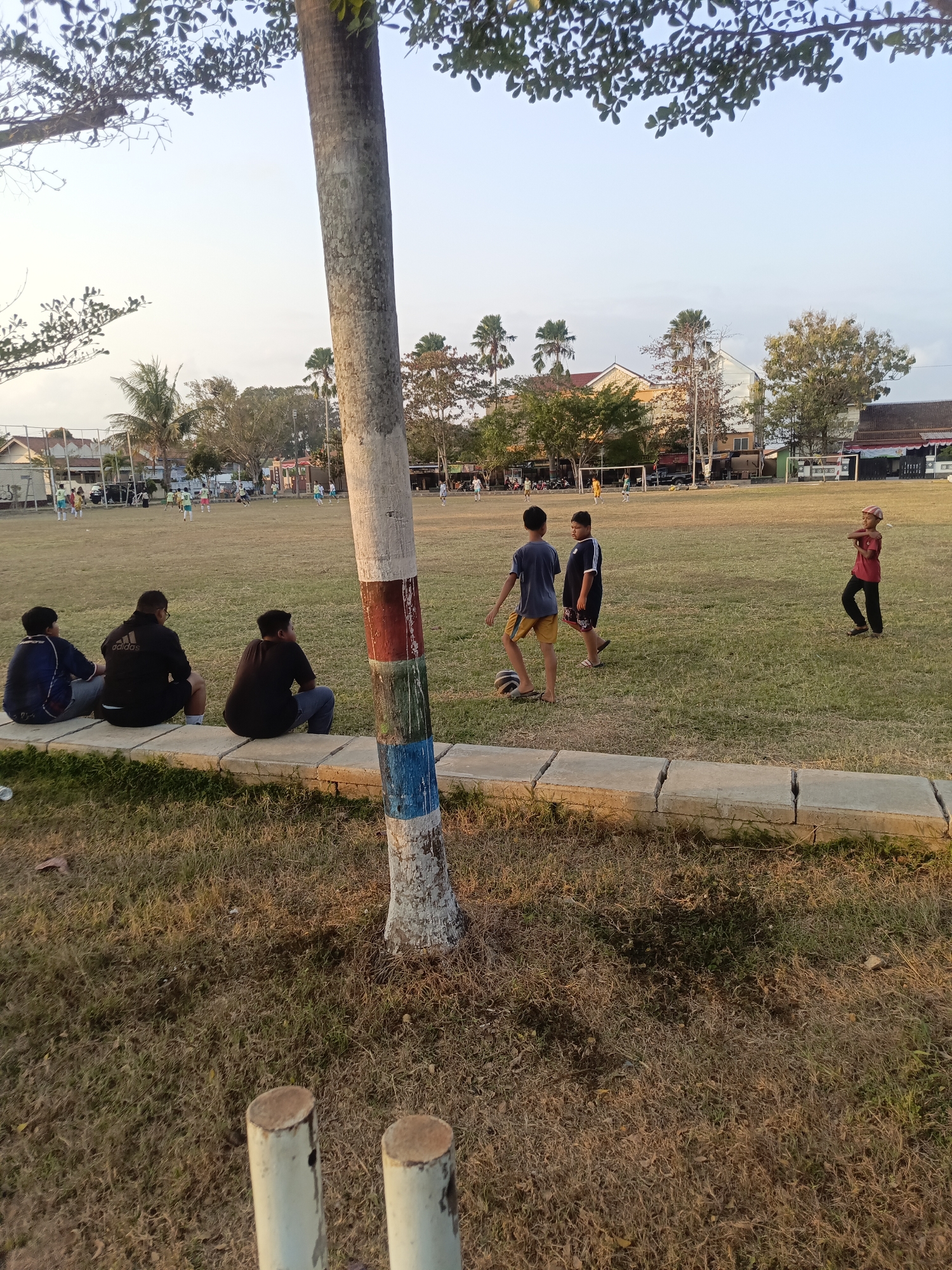 This image depicts a soccer field in Desa Gunungsimping, Indonesia, during daylight hours with a clear sky. In the foreground, three young males are seated on a low concrete barrier, facing away from the camera and towards the field. Their attire consists of casual clothing, including a blue and black jacket, a black jacket with an Adidas logo, and a dark t-shirt. To the right of the seated individuals and on the field, three young males are engaged in a soccer-related activity. One boy, wearing a blue t-shirt and yellow shorts, is near a soccer ball. Another boy, in a black and white t-shirt and black shorts, stands next to him. A third boy, wearing a red t-shirt and black pants, is positioned further to the right. In the middle ground of the field, several other individuals, presumably children, are visible playing soccer, with soccer goals and nets present at either end of the field. The field is covered in dry grass. Behind the field, buildings and trees are visible, indicating a semi-urban or suburban environment. A prominent tree with colorful paint markings on its trunk stands in the right foreground. The overall atmosphere appears to be one of casual recreation and outdoor activity.