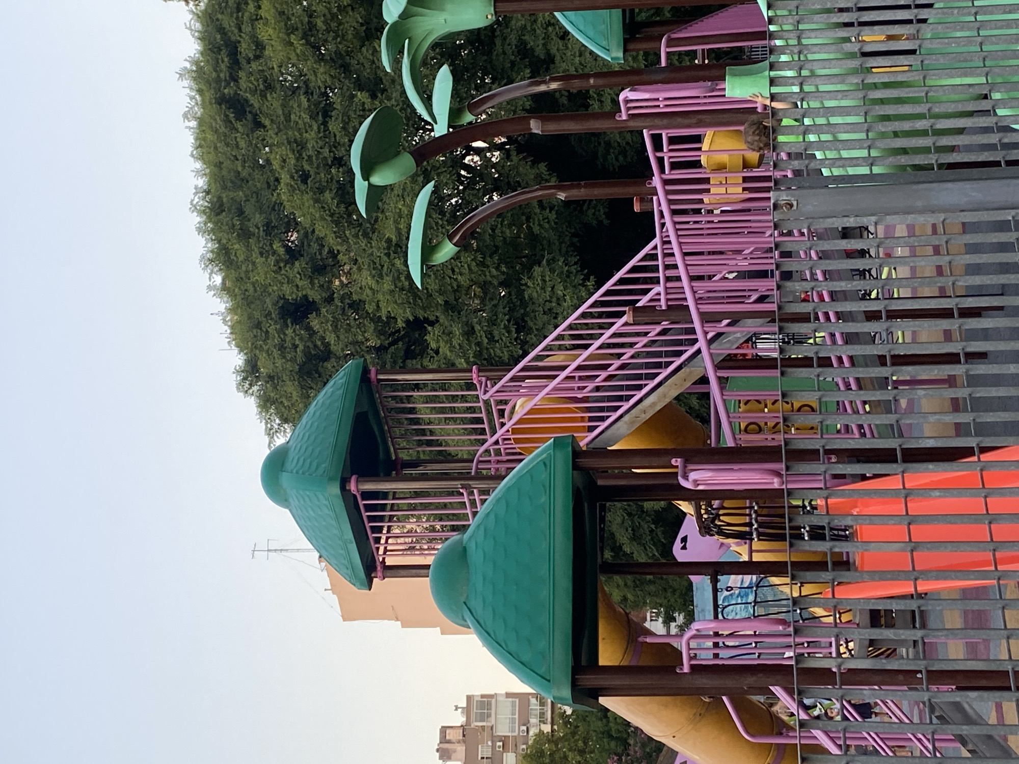 The image shows a playground in Buenos Aires, Argentina. The playground features a climbing structure with green canopies and yellow slides, connected by pink stairs and brown palm tree-shaped decorations. A gray metal fence surrounds the playground. In the background, buildings are visible, as well as lush green trees and a pale sky. A child is seen within the structure.