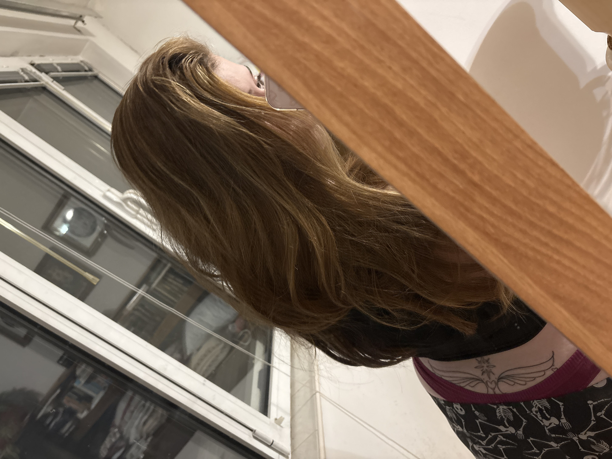 A woman with long brown hair is visible from the chest up, with a partial view of a wooden object in the foreground. She is wearing a black top and patterned leggings. A window with multiple panes and white frames is in the background, showing indistinct shapes and a glimpse of the exterior. The location is not discernible as Troyan, Bulgaria from the image alone.