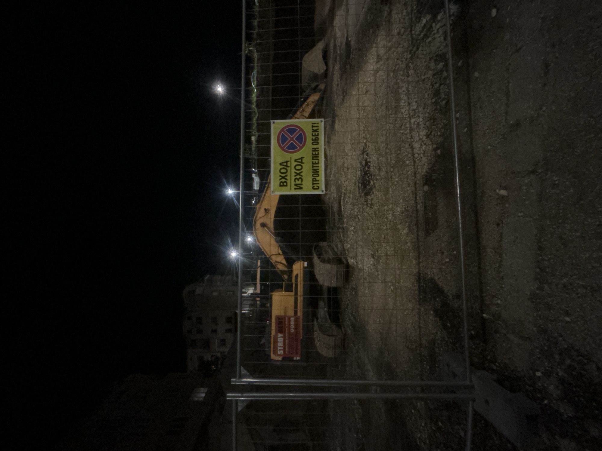 At night in Pleven, Bulgaria, an excavator is behind a metal fence. A sign that says "Construction Object" is attached to the fence.
