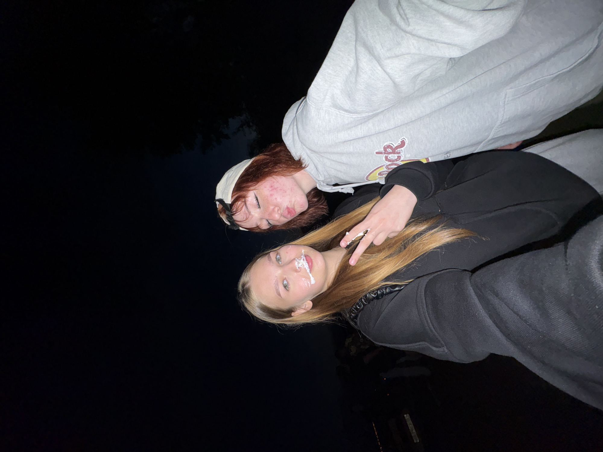 Two young women are posing for a selfie outdoors at night. The woman on the left has long blonde hair and cake frosting on her face, around her mouth and nose. She is wearing a black hooded sweatshirt and is holding up two fingers in a peace sign. The woman on the right has short red hair, a white baseball cap, and a light gray hooded sweatshirt with the "Hard Rock" logo visible. She is puckering her lips, as if for a kiss. The background is dark, with hints of trees and possibly other people, suggesting an outdoor gathering or event. There are no discernible landmarks or visual cues that indicate the location is specifically in the city of Chaikovsky, Russia.
