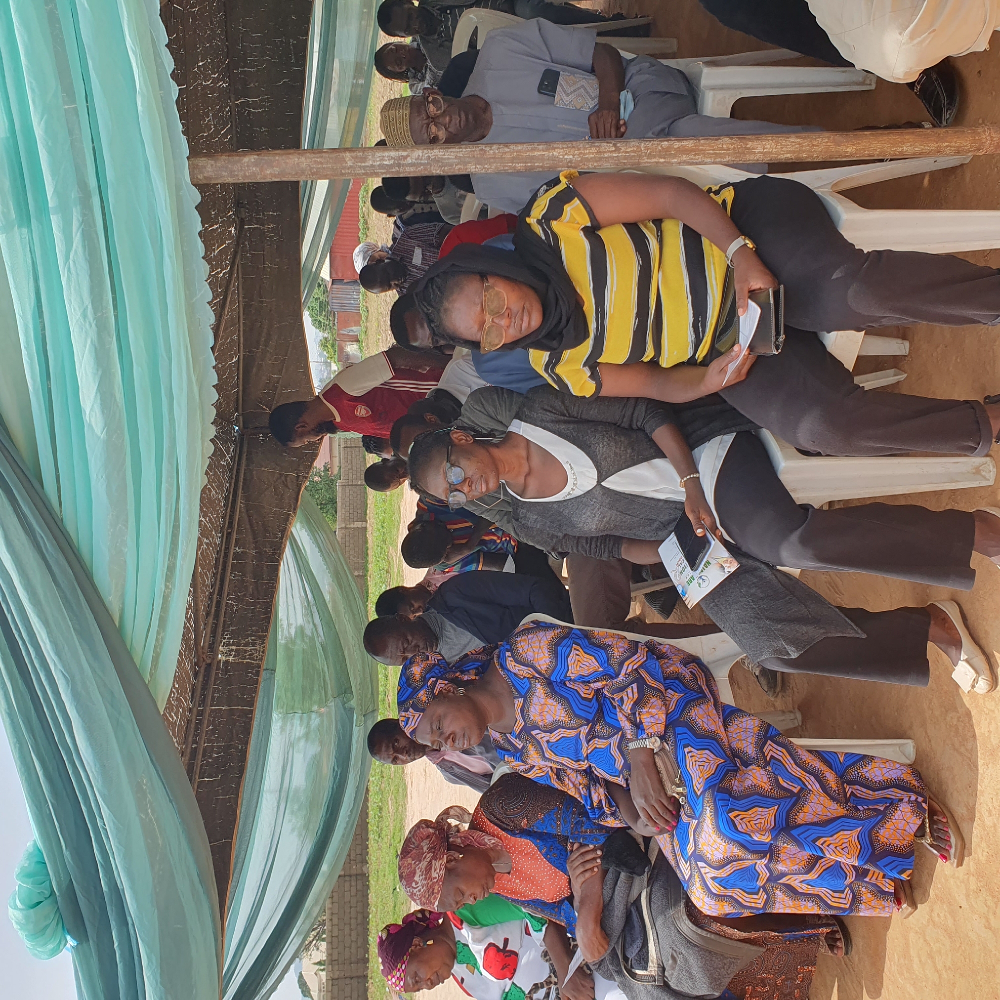 This media file captures an outdoor gathering in Unguwar Saya, Nigeria, likely during the daytime. The scene appears to be a community event or meeting, with a group of people seated on white plastic chairs under a makeshift canopy. The canopy is constructed with a corrugated metal roof covered by brown fabric, supported by wooden beams, and draped with light blue and green fabric curtains.
The subjects are diverse, consisting of men and women of various ages. Many individuals are dressed in traditional Nigerian attire, characterized by vibrant patterns and colors, such as the striking blue and orange geometric print worn by a woman in the foreground. Others are in more casual Western-style clothing. Some people are looking forward, suggesting they are attending to a speaker or presentation, while others have their eyes closed or are looking down, possibly due to the heat or fatigue.
Notable details include the presence of cell phones, with one woman holding a smartphone and another holding documents. A flyer is visible in the foreground with some text, but it is not clearly legible. The ground is covered in reddish-brown earth, and in the background, brick structures and some greenery can be seen, hinting at a rural or semi-urban setting. The overall mood seems to be one of community engagement, with a sense of focus among the attendees. The time of day appears to be late morning or afternoon, given the sunlight and shadows. The weather seems to be warm, as indicated by the attire and the need for shade.