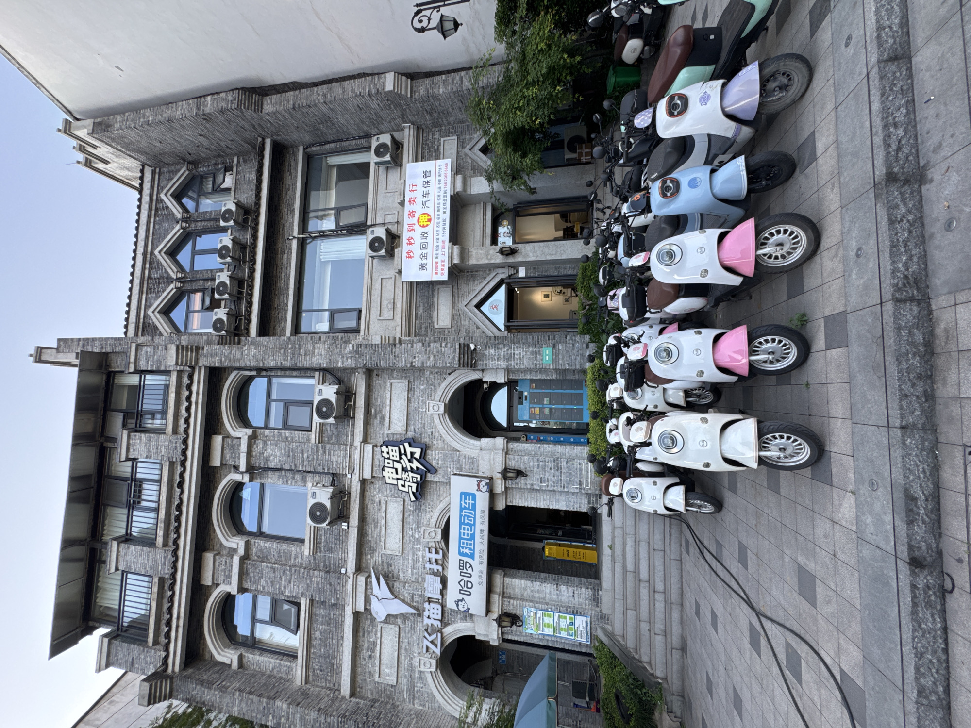 The image depicts a three-story building in the Yuzhong District of China. The building is made of gray brick and has several arched windows and air conditioning units. There are multiple signs in Chinese characters, advertising businesses such as "Flying Cat Motorcycle" and "Hello Rental Electric Vehicle," indicating electric scooter rentals.
Parked alongside the building is a row of electric scooters. They are of varying colors including white, pink, blue, green, and brown.