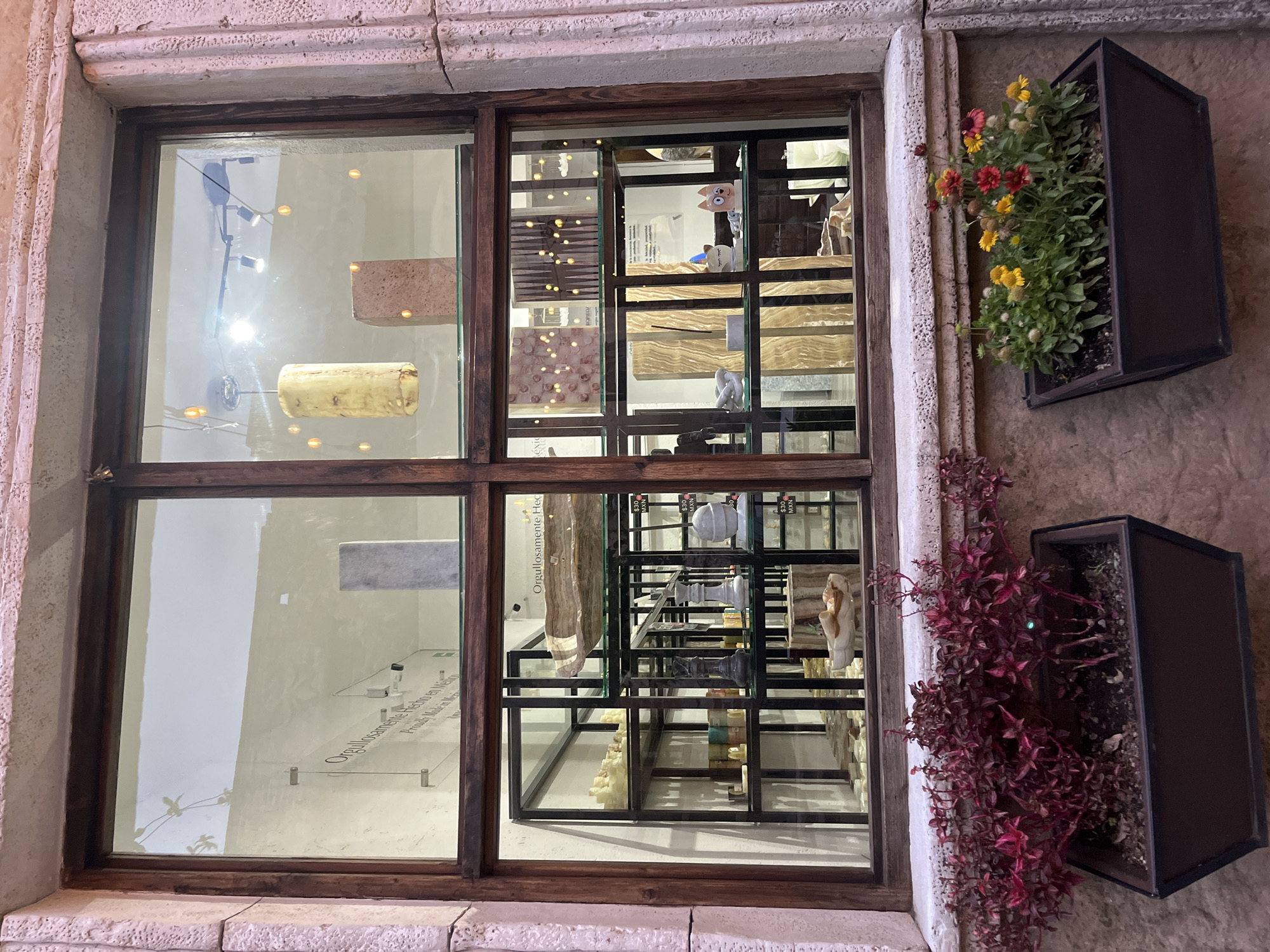 The image shows a window display in Domingo Arenas, Mexico. The window is divided into panes, with the left panes displaying stone slabs and light fixtures. The right panes show a shelf system displaying various stone pieces, including a cartoon cat figurine, and price tags indicating "$30 MXN." Plants are displayed in planters outside the window on the right, with red and green foliage. The wall surrounding the window is textured. The OCR in the window displays the words "Proudly Made in Mexico" and "Orgullosamente Hecho."