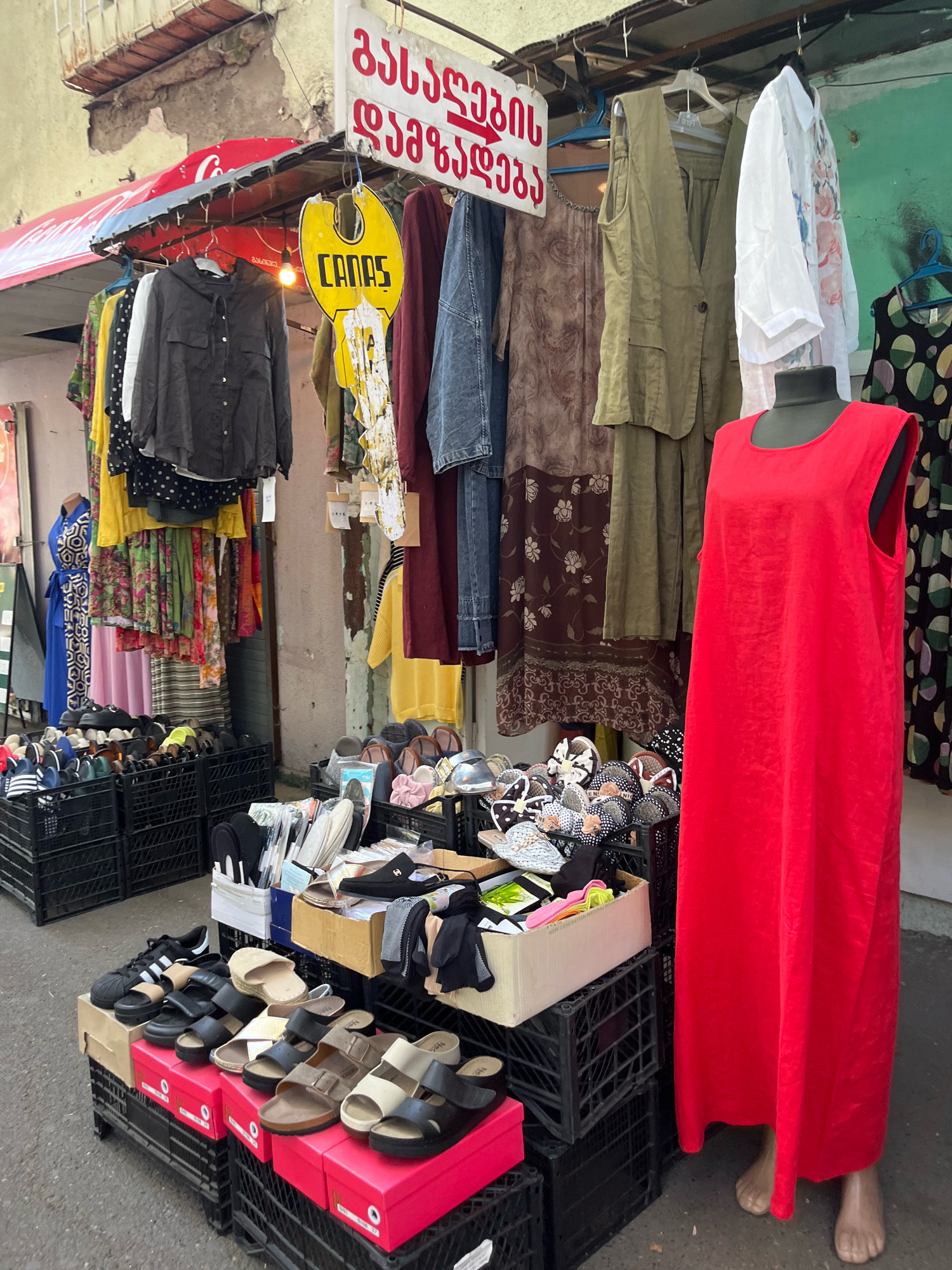 This image depicts a street market stall in Tbilisi, Georgia, displaying a variety of clothing and footwear. The stall features clothing items such as dresses, shirts, and vests hanging from a rack, with a red mannequin in the foreground wearing a red dress. Below the clothing, various types of sandals and flip-flops are arranged in black plastic crates and on top of pink boxes. A yellow sign with the word "CANAS" in black lettering is prominently displayed. The background shows a weathered building facade and a red awning with a Coca-Cola logo. The scene appears to be an outdoor retail environment.