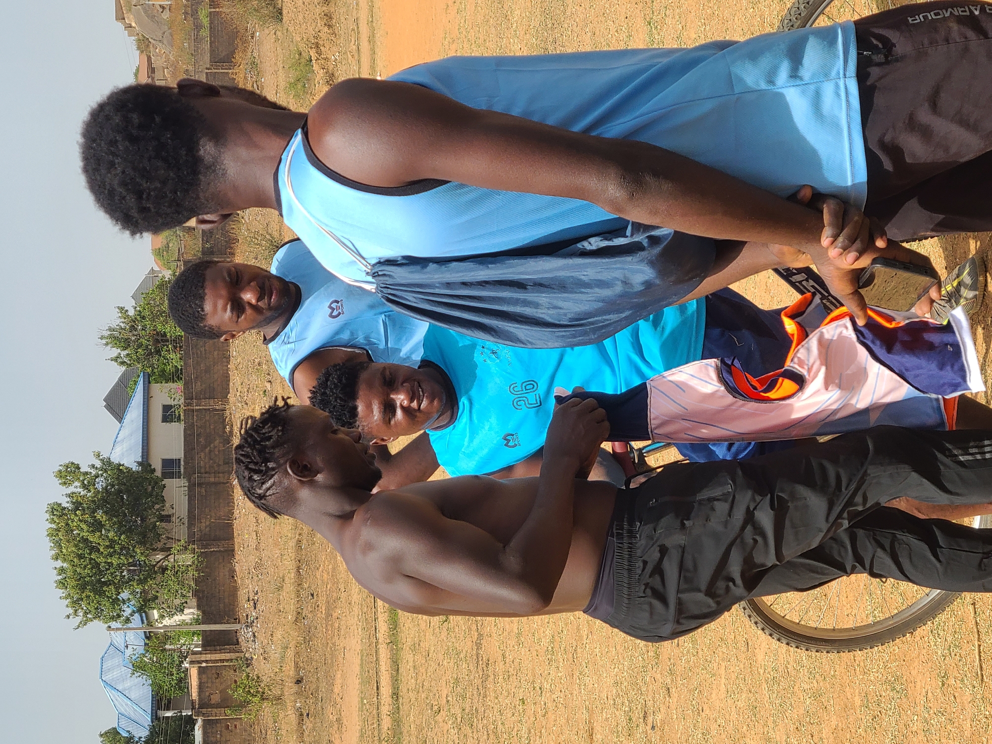 Four men are gathered outdoors in a field near Barikin Jirga Summa, Nigeria. One man stands shirtless and wears black shorts. Another man wears a light blue shirt and black shorts labeled "UNDER ARMOUR". The other two men wear different light blue athletic shirts. One man is holding a phone in his hand and they are holding jerseys. A bicycle wheel is visible near the shirtless man. The ground appears to be dry and dusty, and there are a few trees and buildings in the background.