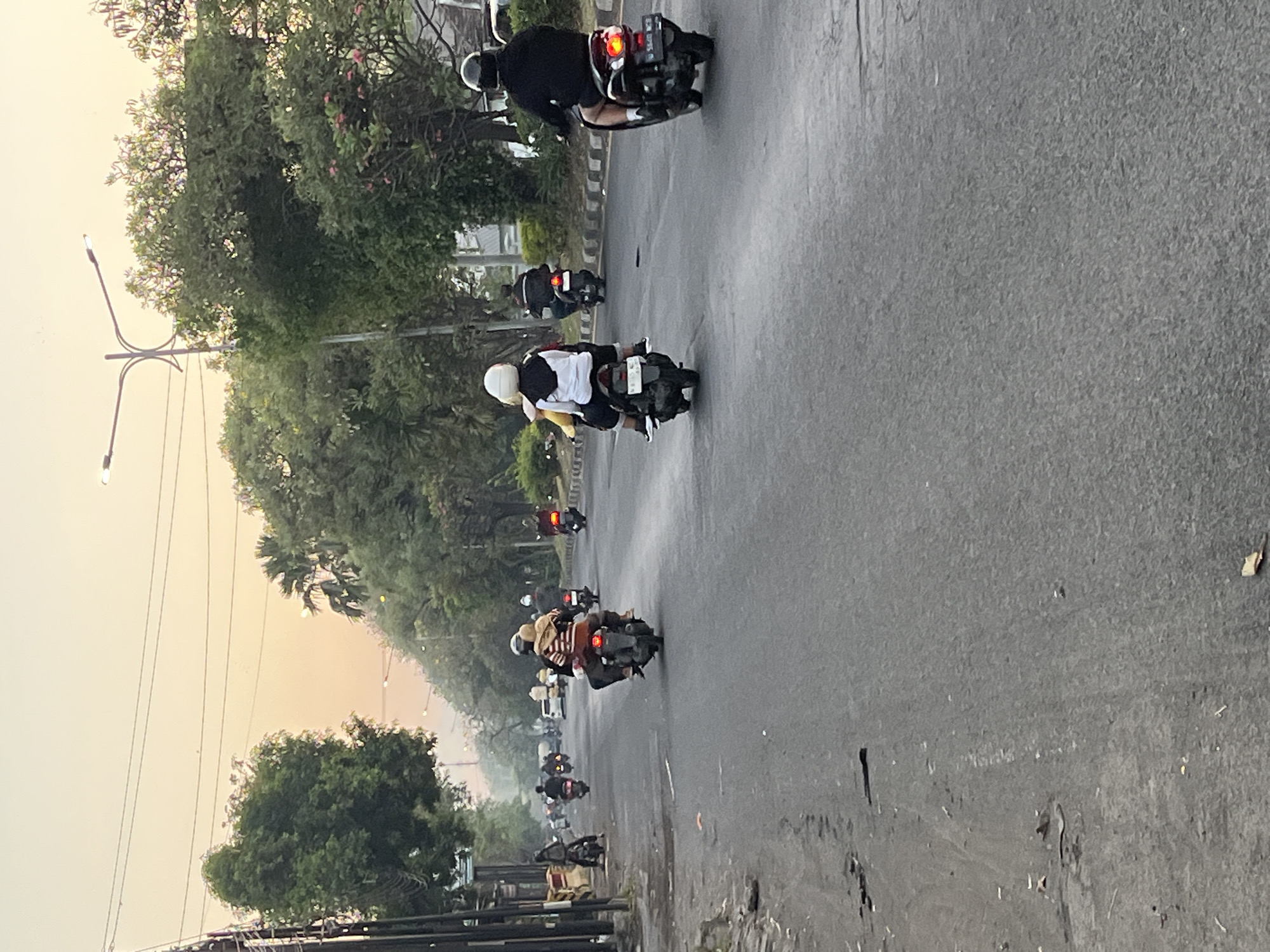 The image depicts a street scene in Ngaban, Indonesia, characterized by a light-colored sky suggesting either dawn or dusk. Several individuals are riding motorbikes along a paved road, which appears to be a city street with trees and vegetation lining one side. The road surface is dark asphalt, with some debris and markings visible. On the left side of the road, there is a sidewalk area with some structures and more trees. On the right side, there are more trees and a curb. The overall impression is that of a typical urban environment with motor vehicle traffic.
Specific details include:
- **Sky:** The sky is a light, hazy orange-yellow, indicating either sunrise or sunset.
- **Road:** The road is paved with asphalt and appears to be multi-lane, with traffic moving in the same direction. There are some potholes and debris on the road surface, particularly on the left side.
- **Vegetation:** Trees and some flowering bushes are visible on both sides of the road, with some taller palm-like trees in the distance.
- **Vehicles:** Multiple motorbikes are present, each carrying one or two individuals. The motorbikes are various colors, but mostly dark.
- **People:** People are wearing casual clothing and helmets for safety. Some are riding alone, while others are carrying a passenger.
- **Infrastructure:** A street light on a pole is visible in the upper right portion of the image. Power lines are also visible overhead.
The scene appears to be a normal, everyday occurrence in Ngaban, Indonesia, with people commuting or traveling on their motorbikes. The time of day appears to be either early morning or late afternoon, judging by the sky color and the lighting conditions.
