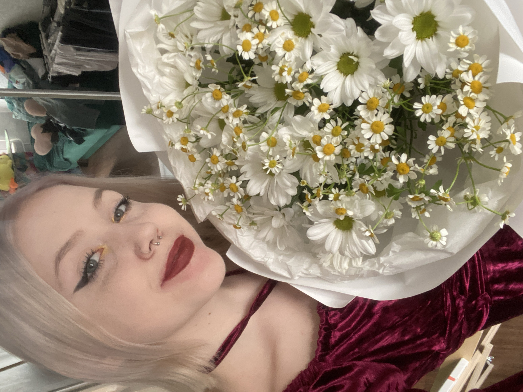 A close-up, tilted portrait shows a young individual with light blonde hair, dark winged eyeliner, yellow eyeshadow, and dark red lipstick, gazing towards the viewer. They have a septum piercing and a nostril piercing. A large bouquet of white flowers, primarily daisies and smaller chamomile-like flowers with yellow centers, is prominent in the foreground, partially obscuring the person's right side. The flowers are wrapped in white paper. The individual is wearing a dark red, possibly velvet, top with thin straps. In the out-of-focus background, shelves with various items, including what appear to be stacked papers or clothes, are visible, suggesting an indoor setting. The individual has a subtle, pleasant expression. The location context provided is Izhevsk, Russia, though no specific visual cues within the image directly confirm this. There is no visible text.