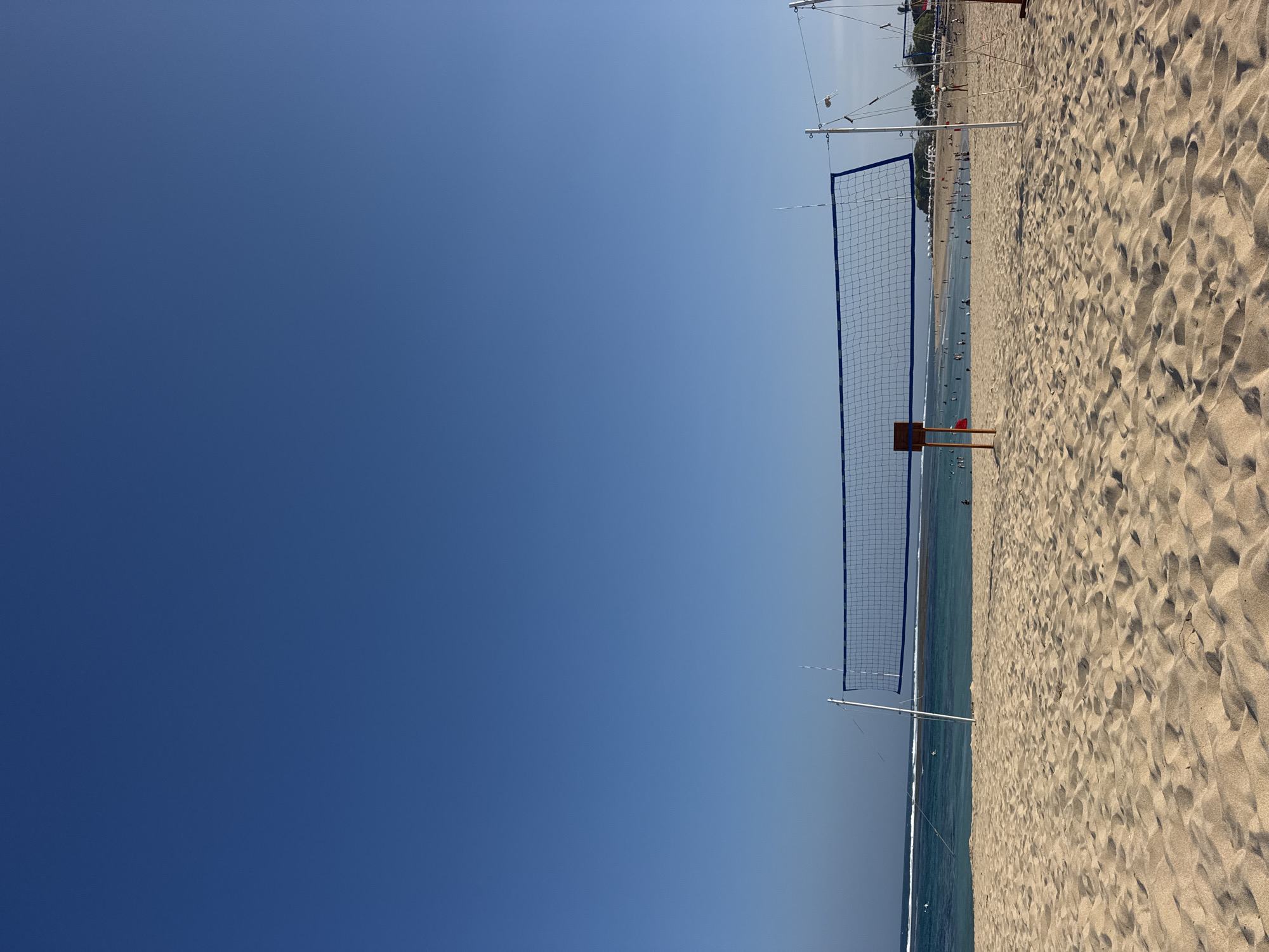 The image depicts a wide expanse of golden sand on Kuta Selatan beach, Indonesia, under a clear, bright blue sky. In the foreground, the sand is textured with ripples from the wind and tide, showcasing the natural beauty of the beach. Dominating the midground is a volleyball net, set up for a game, with its blue netting and white boundary ropes clearly visible. Two wooden posts support the net, one on the left side with an orange marker, and another on the right, partially obscured by other beach structures.
Beyond the net, the turquoise ocean stretches out, meeting the horizon under the vast sky. Small waves gently break near the shore. The water appears calm and inviting, with several people visible enjoying the sea, some wading, others swimming. Further in the distance, along the shoreline, a line of beach umbrellas and palm trees can be seen, indicating a developed resort area. A few more volleyball nets are visible in the far distance, suggesting the beach is a popular spot for recreational activities. The overall scene is one of a serene, sunny day at a tropical beach, with a focus on the recreational aspect of the location.