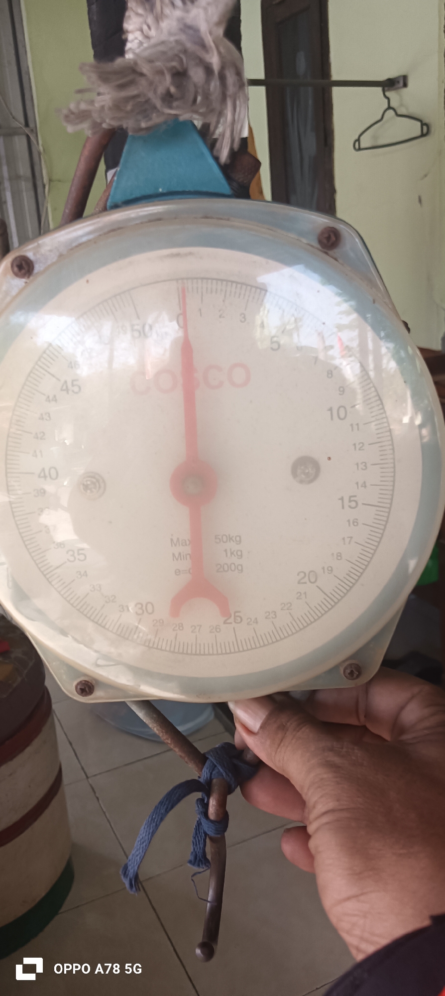 An overhead, close-up, eye-level shot captures a vintage analog scale with a bright red pointer. The scale is circular with a white face and a light blue rim. Markings on the scale indicate weights in kilograms, ranging from 0 to 50. The words "COSCO" are visible on the face of the scale. The pointer is currently positioned between 1 and 2 on the scale. A small tag near the center reads "Max 50kg, Min 1kg, e=200g". A person's hand is visible in the lower right corner of the frame, holding a metal rod that is tied with a blue cord. The background is out of focus but appears to be an indoor setting with a pale green wall and a dark door. The bottom left corner of the image shows a logo that reads "OPPO A78 5G". This image was taken in the city of Buncitan, Indonesia.