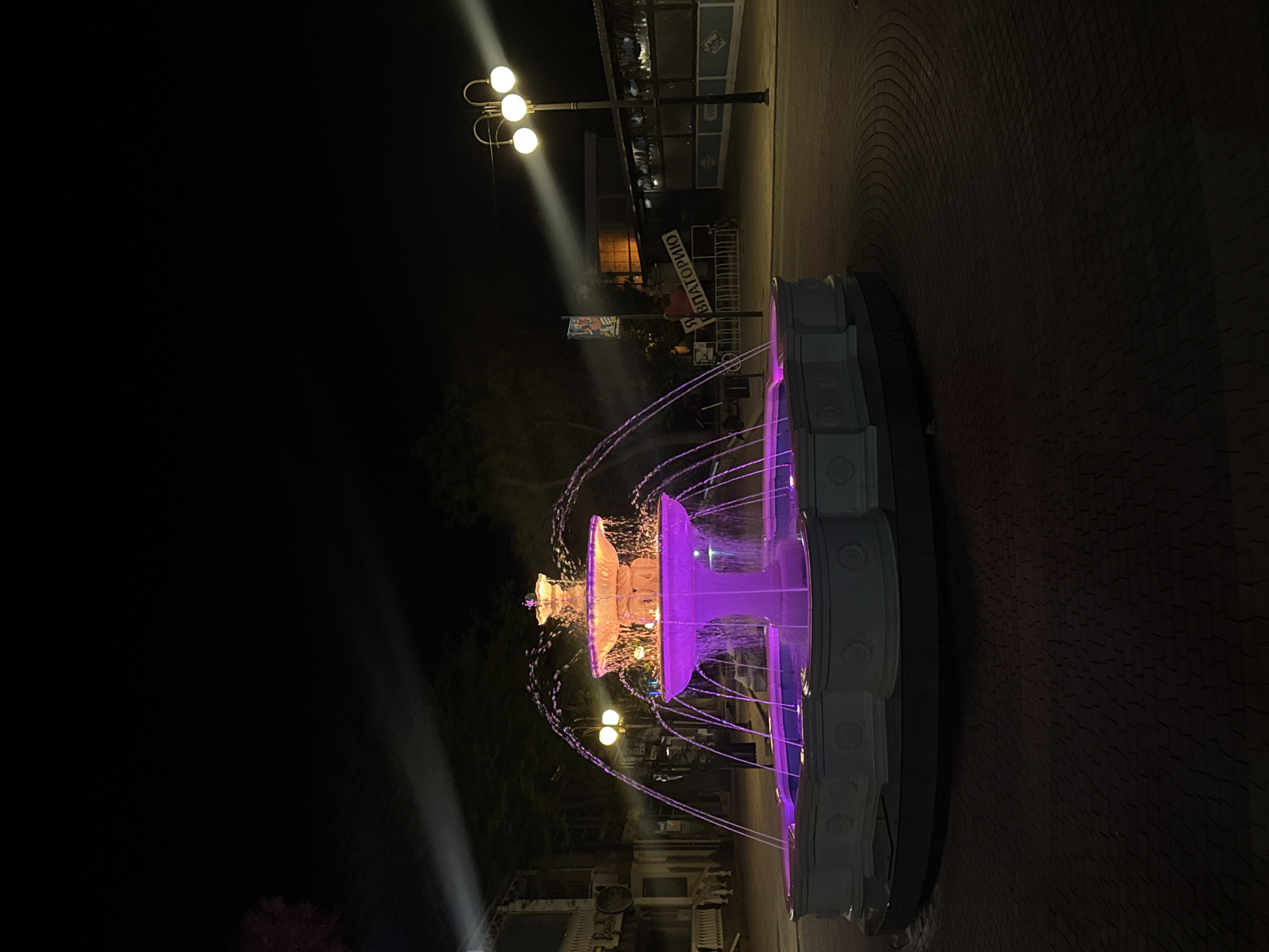 A fountain illuminated with purple and orange lights is positioned on a brick paved area in the city of Yevpatoriya, Ukraine. It is night. A sign in Cyrillic reads "Blagotvorino". Streetlights are visible in the background. The fountain is multi-tiered with water spraying from each tier.