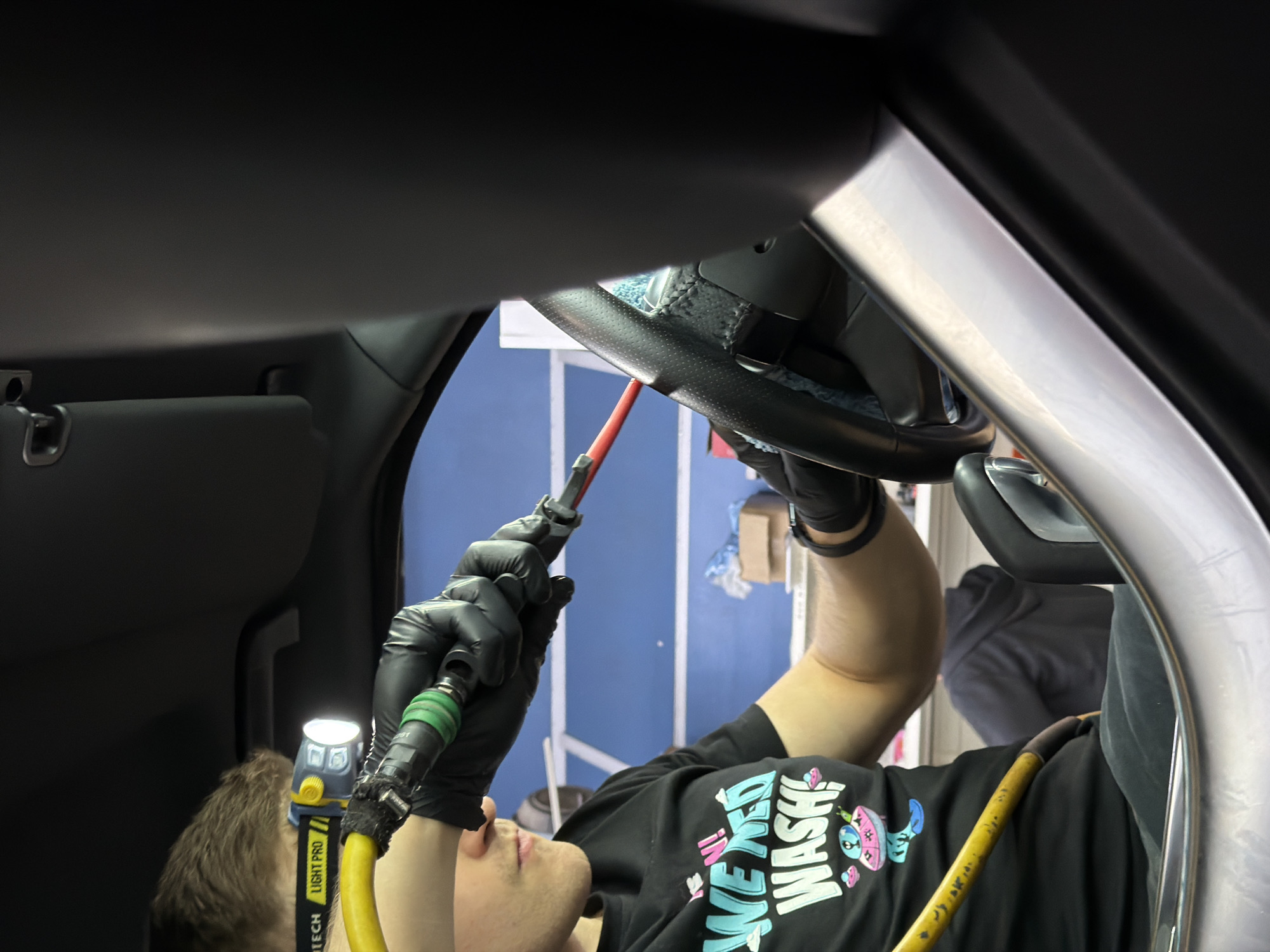 A person wearing a black t-shirt with the text "WEEKEND WASH" and a graphic, black gloves, and a "LIGHT PRO" headlamp is cleaning a car's steering wheel with a red nozzle attached to a yellow hose. The interior of the car is visible, including the dashboard and passenger seat. The exterior background shows a blue wall and white scaffolding, suggesting a garage or workshop setting. The location within the city of Krasnoyarsk, Russia cannot be determined from the visual information provided.