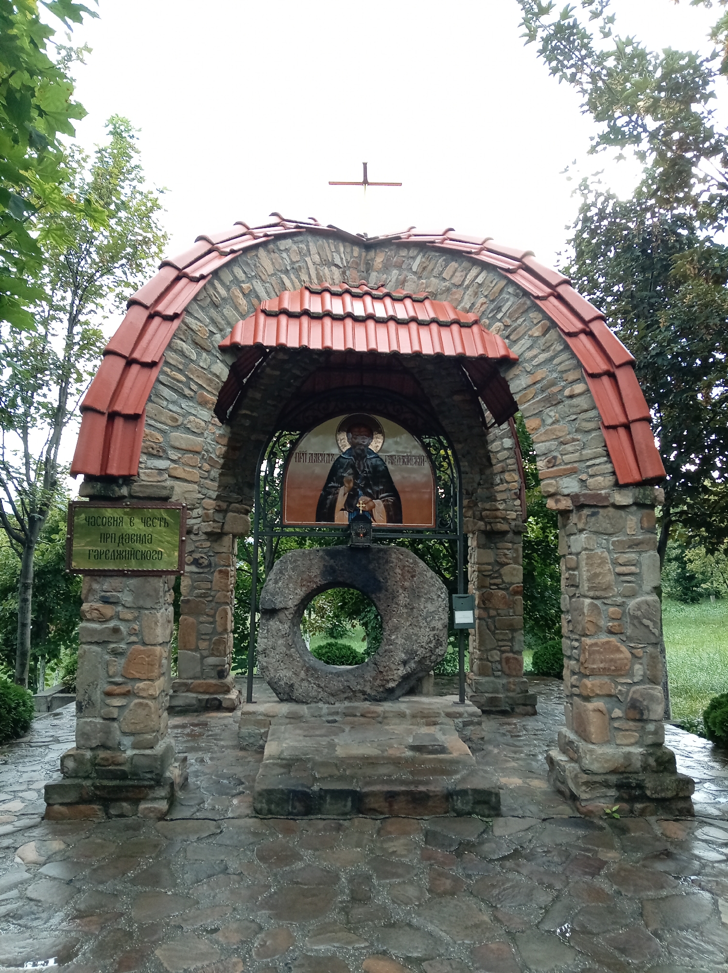 This is a vertical, eye-level, full shot of a stone chapel with a red tile roof, located in the city of Yessentuki, Russia. The chapel is supported by two stone pillars and has an arched entryway. Inside the chapel, there is an icon of a saint and a large, circular stone. The ground in front of the chapel is paved with stones and appears wet. Trees and greenery surround the chapel, indicating it is outdoors.