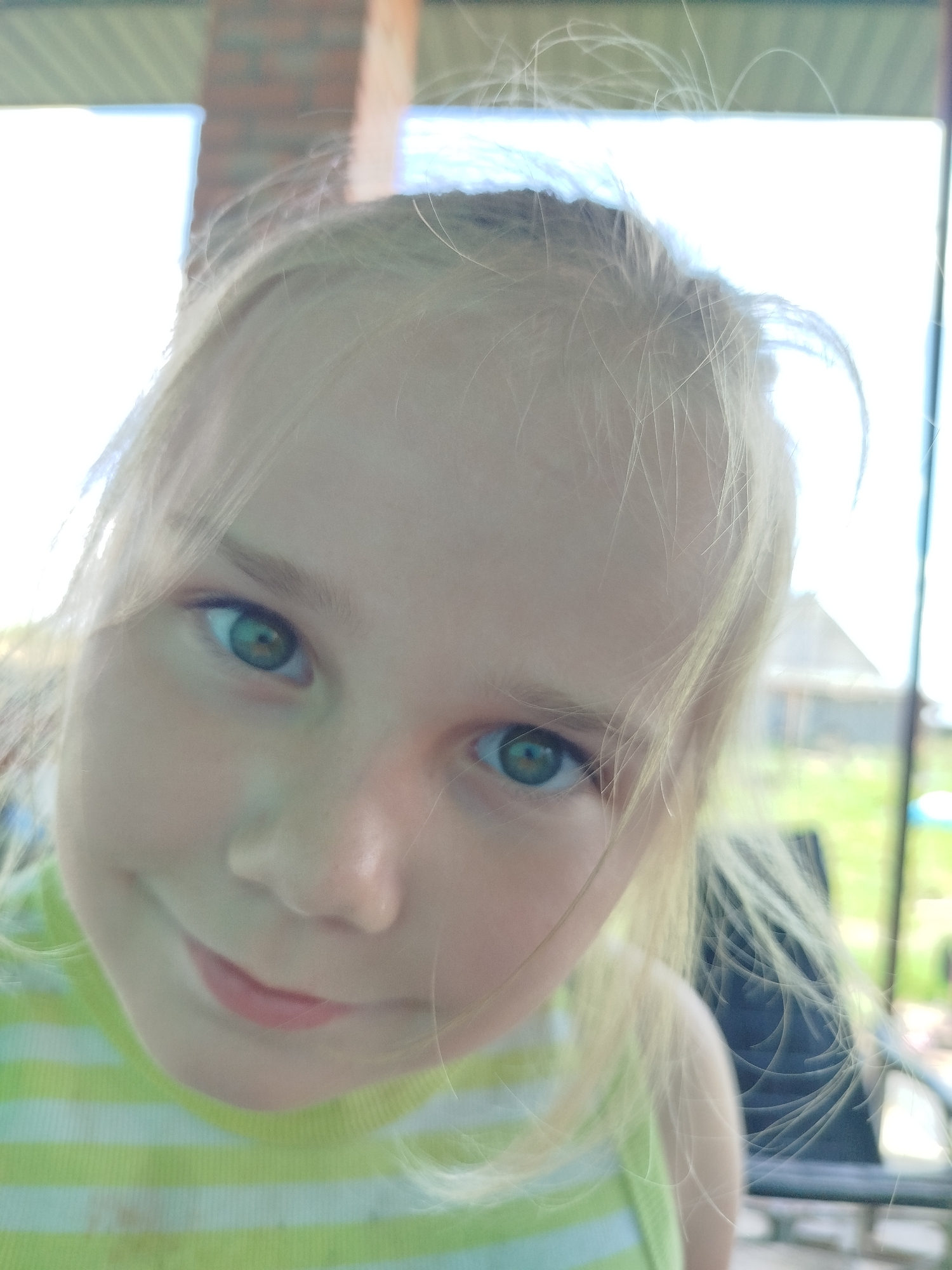 The image is a close-up, candid portrait of a young blonde girl. She is looking directly at the camera with a slight smile. Her eyes are green and appear to have a darker ring around the pupil. Her hair is light blonde and somewhat disheveled, with strands falling across her face. She is wearing a green and white striped t-shirt. The background is blurred, but appears to be an outdoor setting with some structures and greenery, possibly a yard or patio area. The lighting suggests it is daytime. No specific location in Крывасельскі сельскі Савет, Belarus can be determined from the visual information provided.