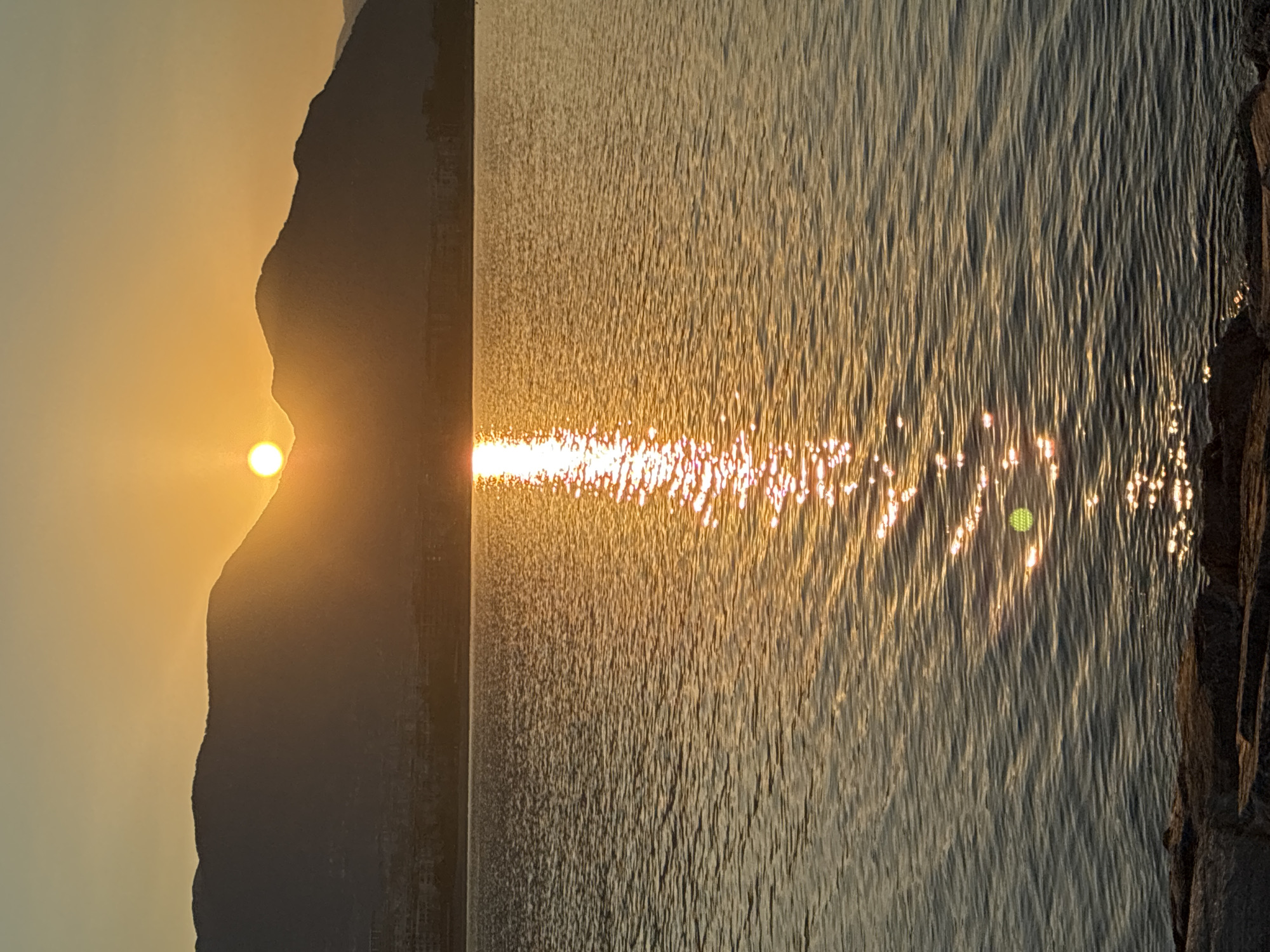 A tranquil scene unfolds as the sun, a luminous orb, dips behind a silhouetted mountain range, casting a radiant golden path across the calm waters of the Mediterranean Sea. The sky transitions from a soft yellow at the horizon to a pale orange and then a muted grey towards the top of the frame, reflecting the gentle descent of daylight. The surface of the sea is gently rippled, creating a shimmering effect as sunlight dances upon it, with the most intense reflections forming a bright, unbroken line that stretches from the horizon towards the foreground. Along the base of the mountains, faint outlines of buildings suggest a coastal town. In the immediate foreground, the rough texture of large, dark stones lining the shore provides a grounded contrast to the ethereal glow of the sunset. The image captures the serene beauty of Alanya, Turkey, at dusk.