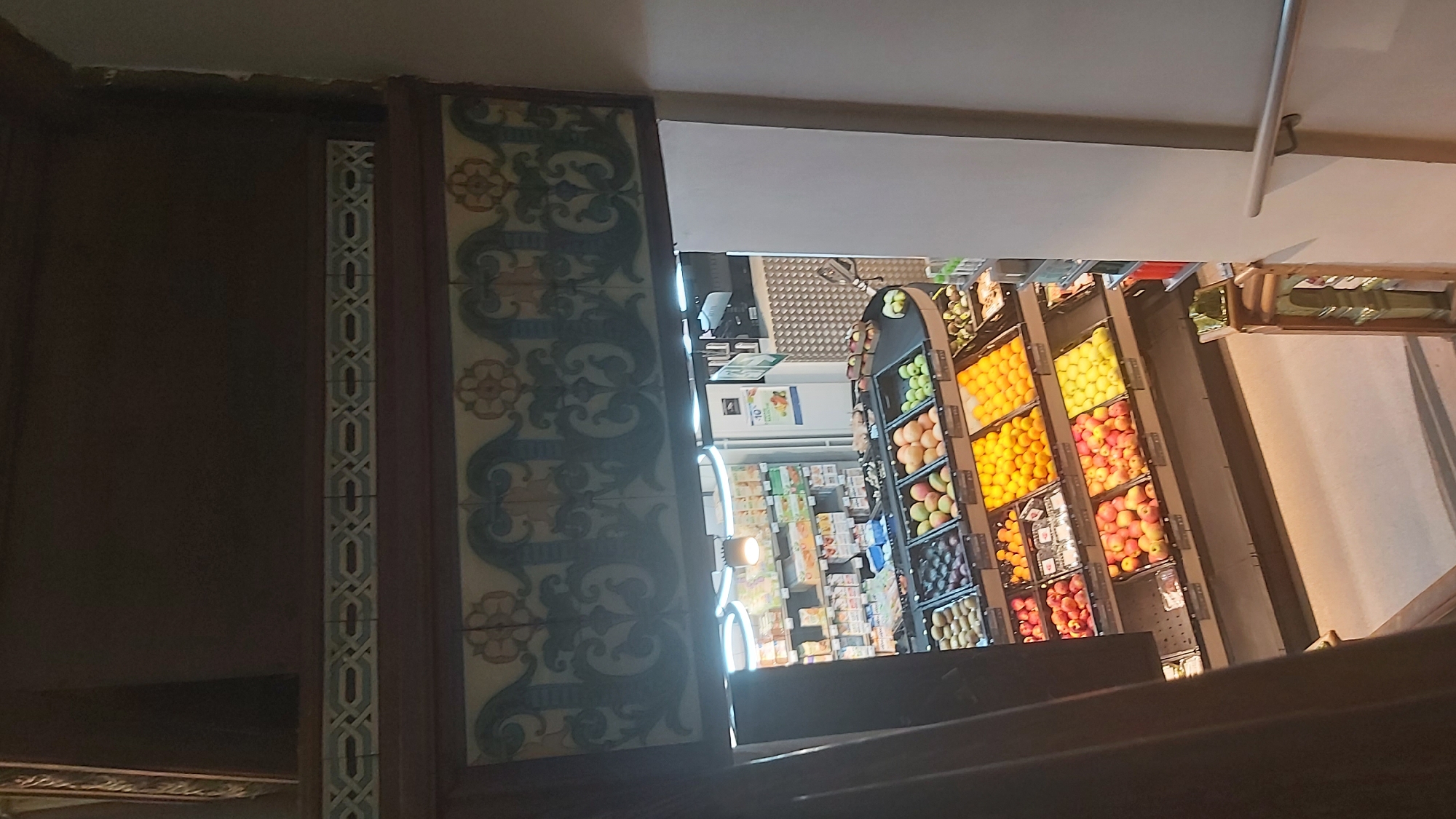 The image shows a view looking down into a grocery store, likely a smaller, specialty market, from an elevated position, possibly a staircase or balcony. The store is well-stocked with a variety of fresh produce displayed in tiered shelves. Fruits such as apples, oranges, and what appear to be mangoes and plums are visible. The store interior features overhead lighting and shelving units containing packaged goods. The decorative architectural elements above the store entrance, including patterned tiles and ornate moldings, suggest a historic building, possibly in Paris, France. The overall impression is one of a clean, organized, and inviting retail space.