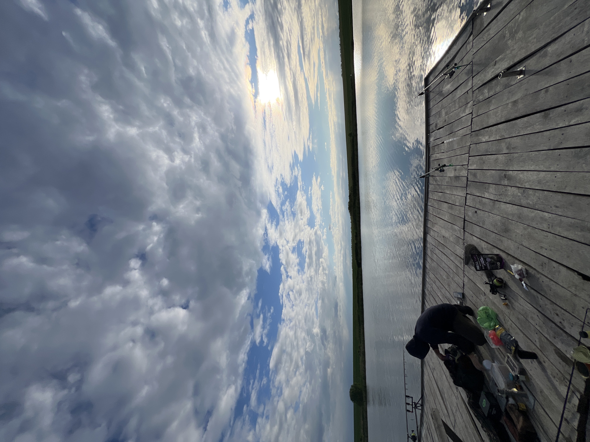 The image depicts a scene at a body of water, likely a lake or pond, in Kopychyntsi, Ukraine. A wooden dock extends into the water. A man is crouched on the dock, possibly preparing fishing gear. Fishing rods are set up along the dock's edge. The sky is partly cloudy with the sun visible. The water reflects the sky. There is green foliage visible across the water. Fishing equipment and tackle are scattered around the man on the dock.
