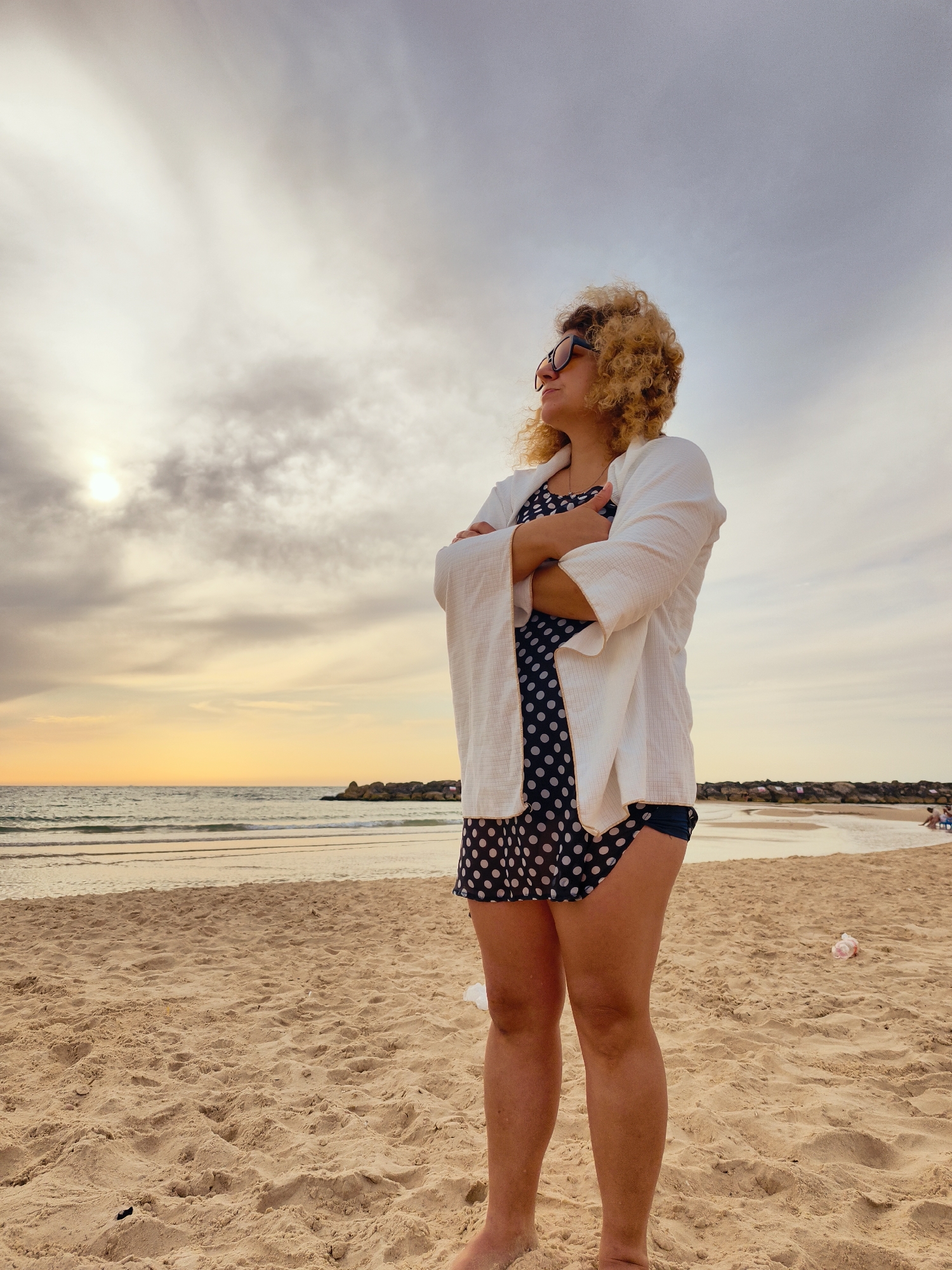 The image displays a serene beach scene, likely at sunset, featuring a woman as the primary subject. The setting is a sandy beach next to the sea in Ashkelon, Israel.
The woman stands prominently in the center-right of the frame, facing left and looking towards the horizon. She has curly blonde hair and wears sunglasses. Her attire consists of a white, long-sleeved top or jacket worn over a dark blue polka-dot dress or swimsuit, with dark shorts visible underneath. Her arms are crossed, conveying a sense of contemplation or calm observation.
The sky occupies the upper portion of the image, characterized by a mix of bright, illuminated clouds near the sun's position on the left, and darker, scattered clouds across the top. The sun, partially obscured by clouds, casts a warm, golden glow across the sky and the water, indicating either sunrise or sunset. The sea in the mid-ground shows gentle waves lapping at the shore. Towards the right side of the water, man-made rock barriers or groynes are visible, extending into the ocean. Distant figures of other people can be faintly discerned further along the beach to the far right. The foreground is a sandy expanse with footprints and a few scattered pieces of light-colored litter.
The overall atmosphere is tranquil and reflective. No visible text is present in the image.