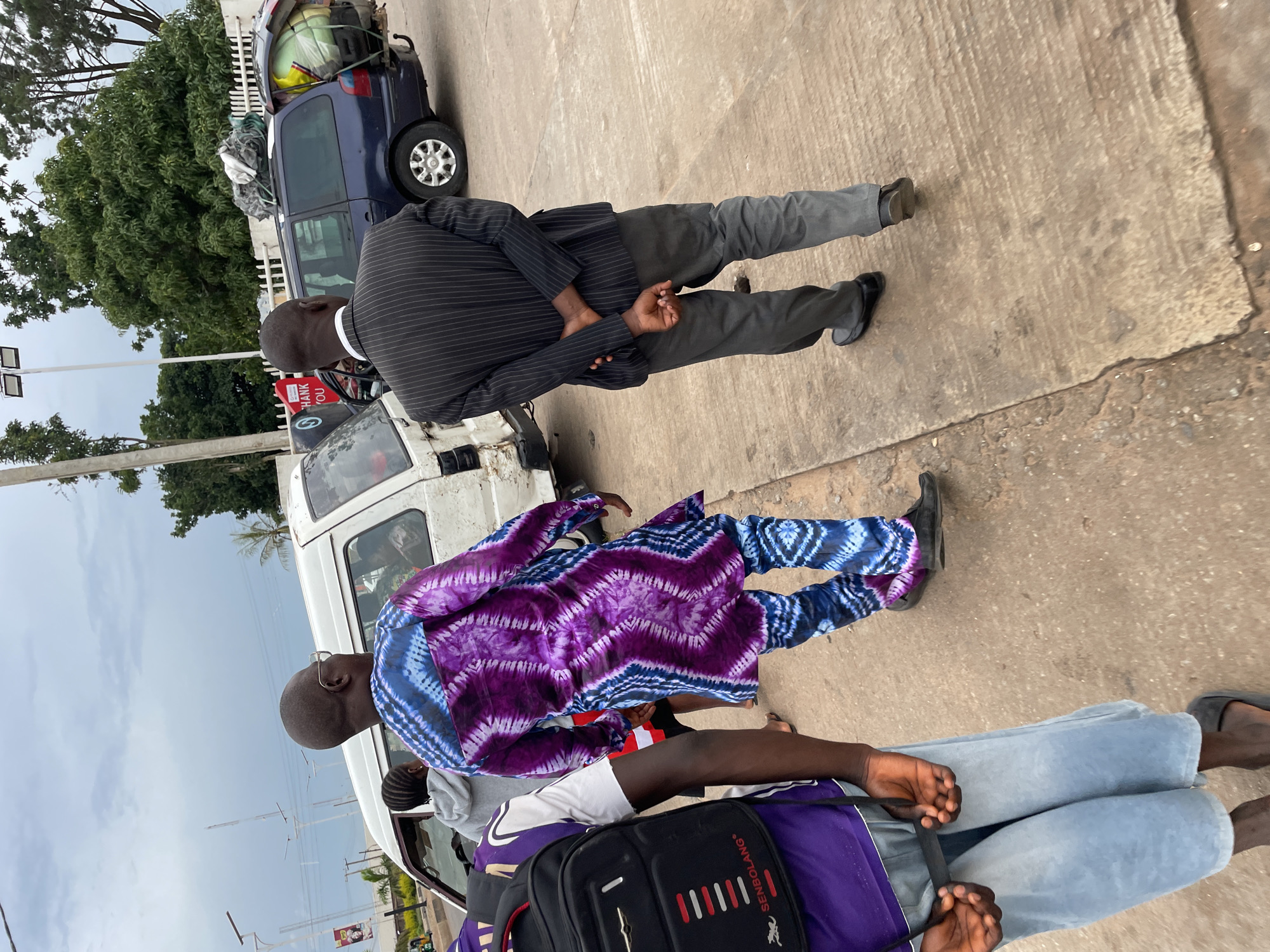In Jos, Nigeria, on a cloudy day, a man in a striking purple and blue tie-dye outfit walks with his hands behind his back, his gaze directed forward. Beside him, another man in a pinstripe suit and grey pants also walks with his hands clasped behind him. A white van is parked behind the man in tie-dye, and a dark blue minivan with luggage strapped to its roof is parked to the right of the man in the suit. The scene takes place on a paved road, with trees and streetlights visible in the background.
