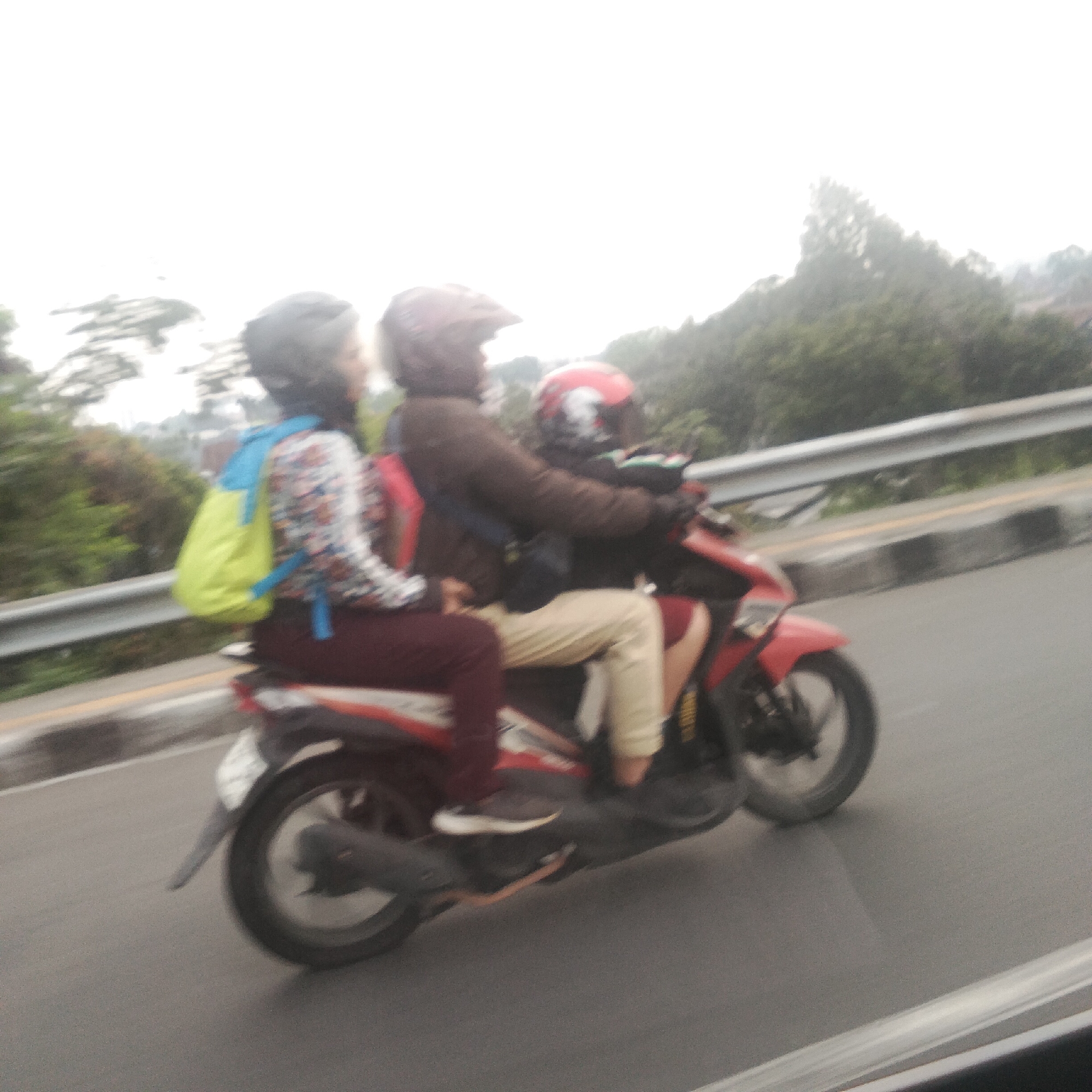 This image shows three people riding on a red scooter on a road in Bogor, Indonesia. The scooter is traveling from left to right. The person in the front is wearing a brown jacket and a helmet. The person in the middle is wearing a patterned shirt and a blue backpack. The person in the back is wearing a red helmet. The road is paved and has a guardrail on the right side. There are trees and foliage in the background. The sky is overcast.