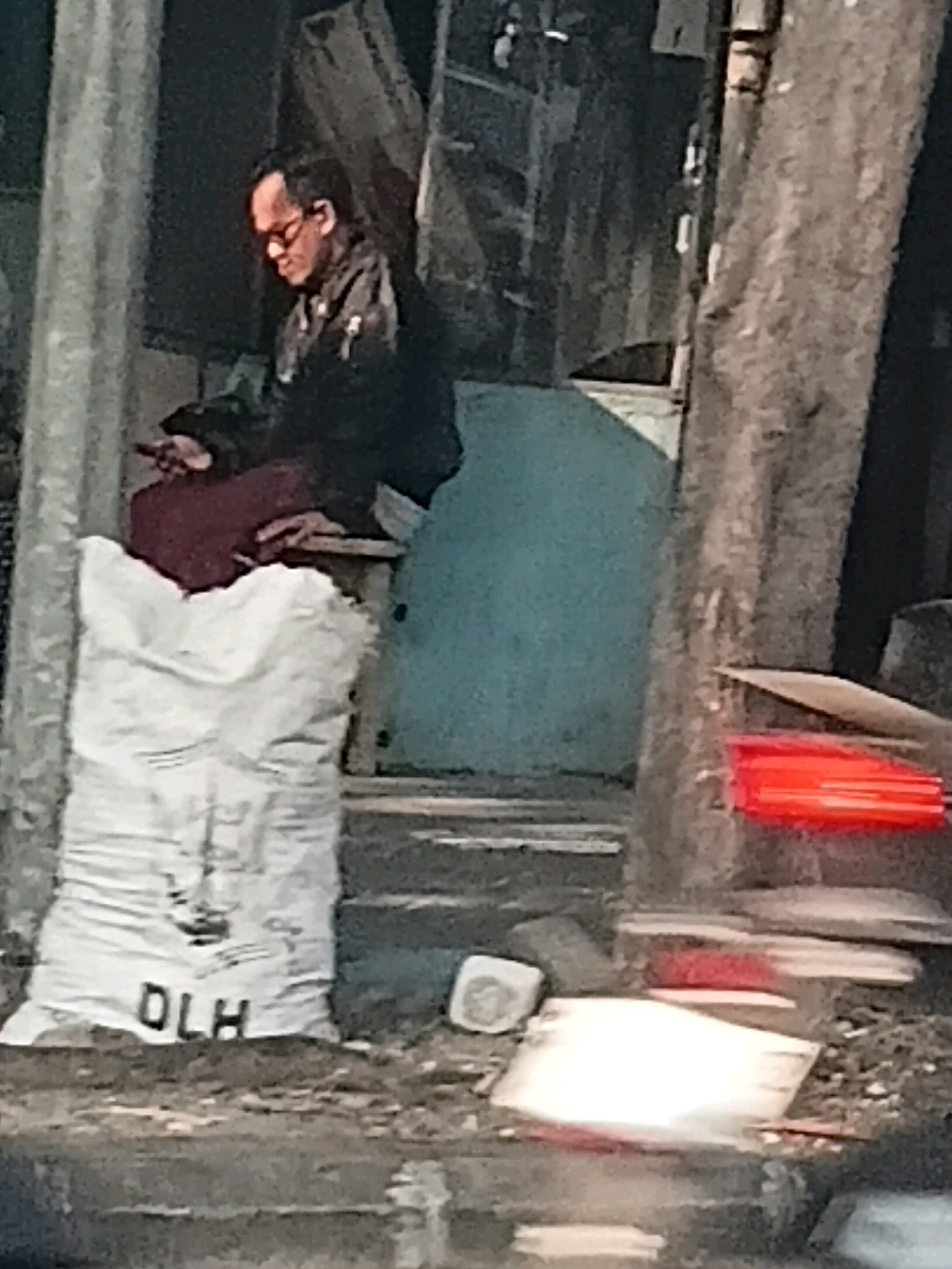 A man wearing glasses and a dark jacket is seated on a wooden stool next to a large white sack. He appears to be holding a cigarette. The sack has "DLH" printed on it. The scene is outdoors, with a rough concrete pillar on the left and a textured, possibly wooden, vertical structure on the right. In the background, there is a blue wall and various discarded objects. There are also some red objects in the lower right quadrant of the image. The overall impression is of a candid, somewhat gritty urban environment.