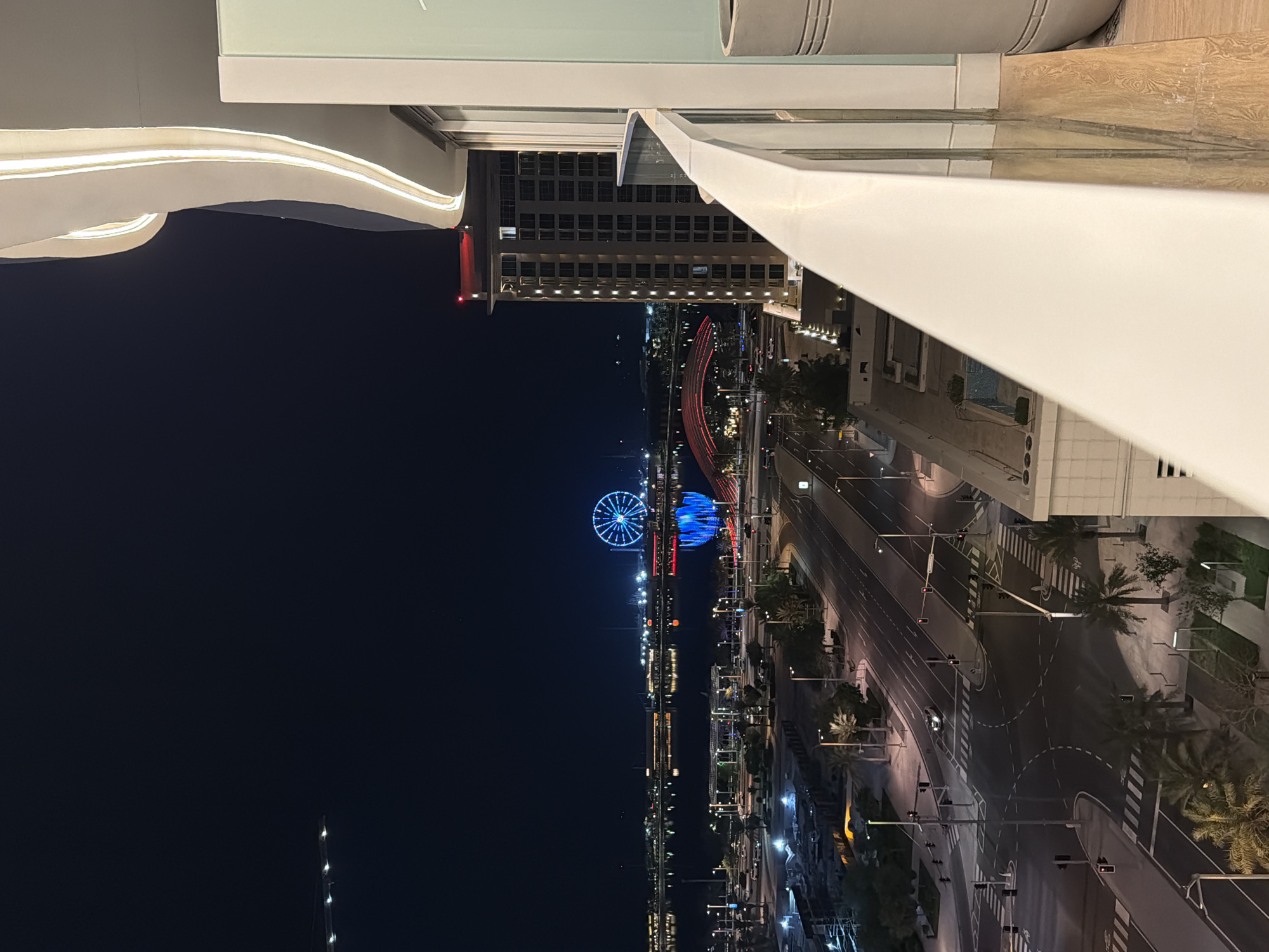 A nighttime, high-angle, wide shot captures a city scene from a balcony. In the foreground, a white railing curves into the frame, with a section of a building and a large planter to its right. The midground shows a street intersection with traffic lights and crosswalks, with cars driving through. Palm trees are visible along the street. In the background, across a body of water, a brightly lit Ferris wheel stands out against the dark sky. A red arched bridge is also illuminated, reflecting in the water. A tall building with many lit windows is to the right of the Ferris wheel. The sky is dark blue.