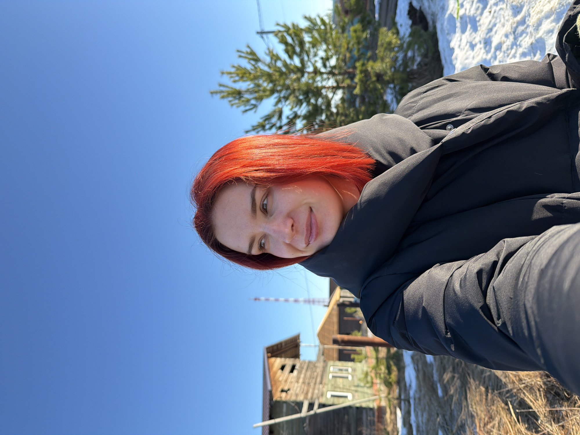 A woman with red hair wearing a black puffy coat is photographed outdoors on a sunny day. The location is identified as the city of Chaikovsky, Russia, based on the presence of a distinctive red and white striped tower in the background. A wooden building with multiple windows and varying degrees of disrepair is also present. A tree and patches of snow are visible in the background. The woman is looking towards the left of the image.