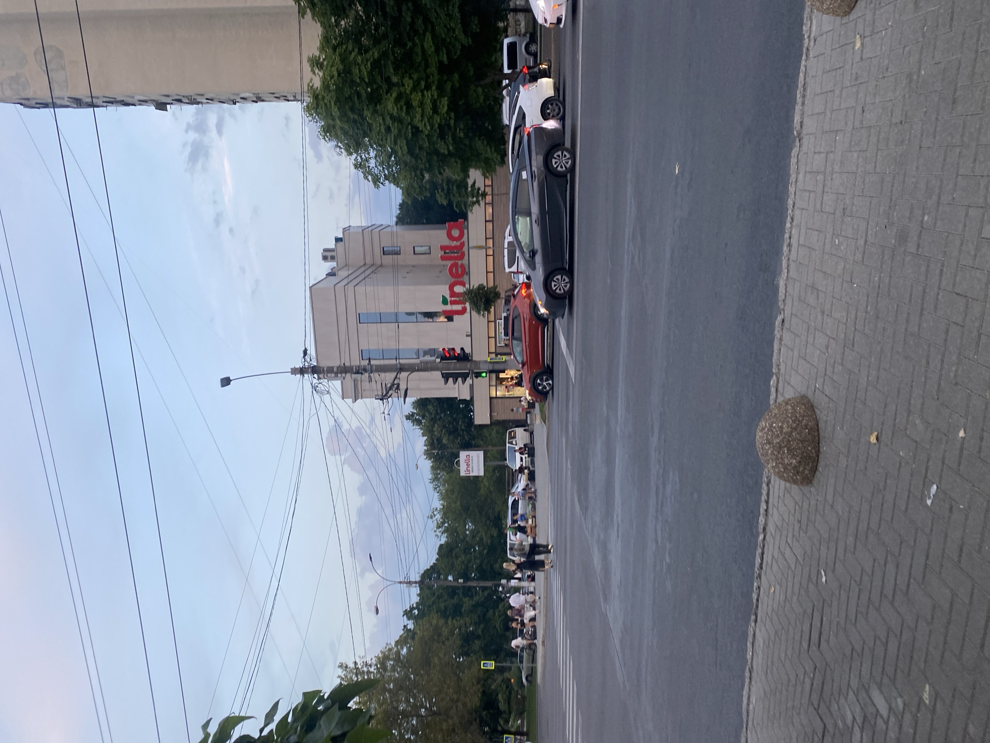 The image depicts a street intersection in Chișinău, Moldova, at dusk. A multi-lane road occupies the foreground, with a pedestrian crossing visible. Several cars are stopped at the intersection, waiting for the traffic light. On the right side of the road, a modern, multi-story building with the "Linella" logo prominently displayed is situated behind trees. The sky is cloudy with a muted, twilight hue. Several power lines crisscross the sky, and a lamppost is visible near the building. People are seen standing and walking on the sidewalk on the left side of the intersection. A decorative stone bollard is placed on the paved sidewalk in the lower right corner of the image.