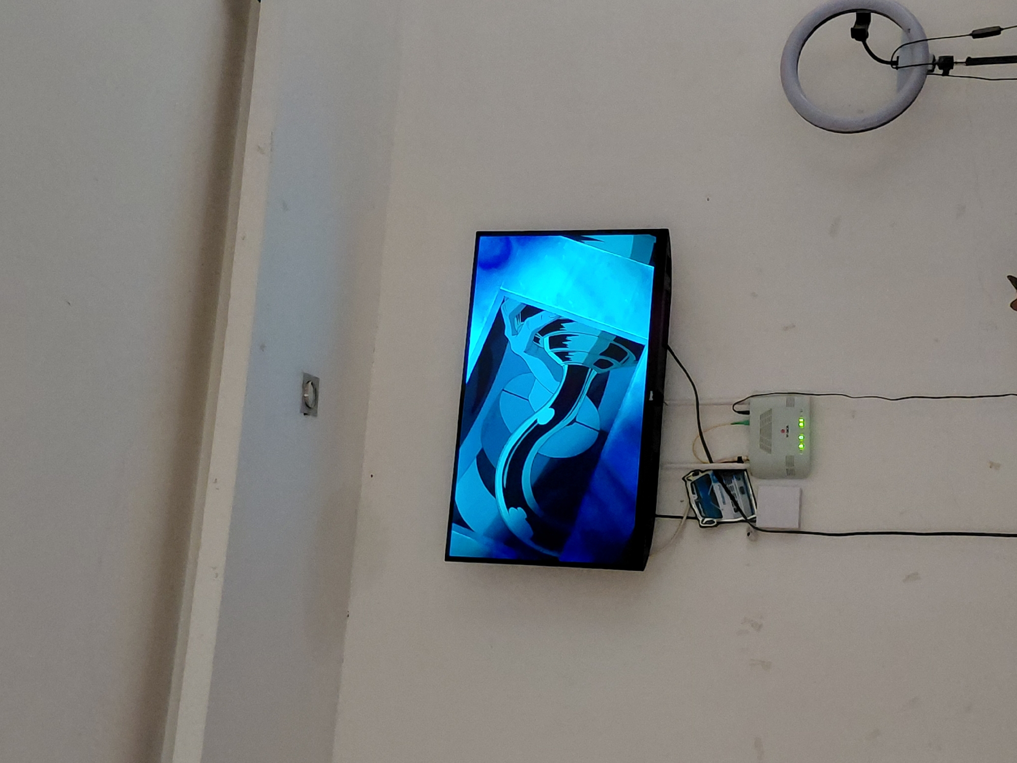 An interior scene in Santa Ana, Venezuela, features a light-colored wall with various electronic equipment mounted on it. A flat-screen television is centrally positioned, displaying an abstract graphic dominated by vibrant blue hues. The image on the screen consists of curved lines and stylized shapes, giving a dynamic, fluid impression.
Above and to the right of the television, a white circular LED ring light is attached to the wall, with black cables extending from it. To the right of the screen, a white network device, likely a modem or router, is mounted. This device has several illuminated green indicator lights. Below it, a smaller white box is also affixed to the wall, and multiple black wires run horizontally across the wall, connecting these devices and extending beyond the frame.
On the far left of the image, a vertical white structural element, possibly a doorframe or part of a wall, runs from top to bottom. A small, metallic, circular fixture is embedded in the wall near this structure. The wall itself is a plain, light color, showing some minor smudges or marks. No people are visible, and no legible text can be discerned on any of the objects. The setup suggests a functional area for media display or communication.