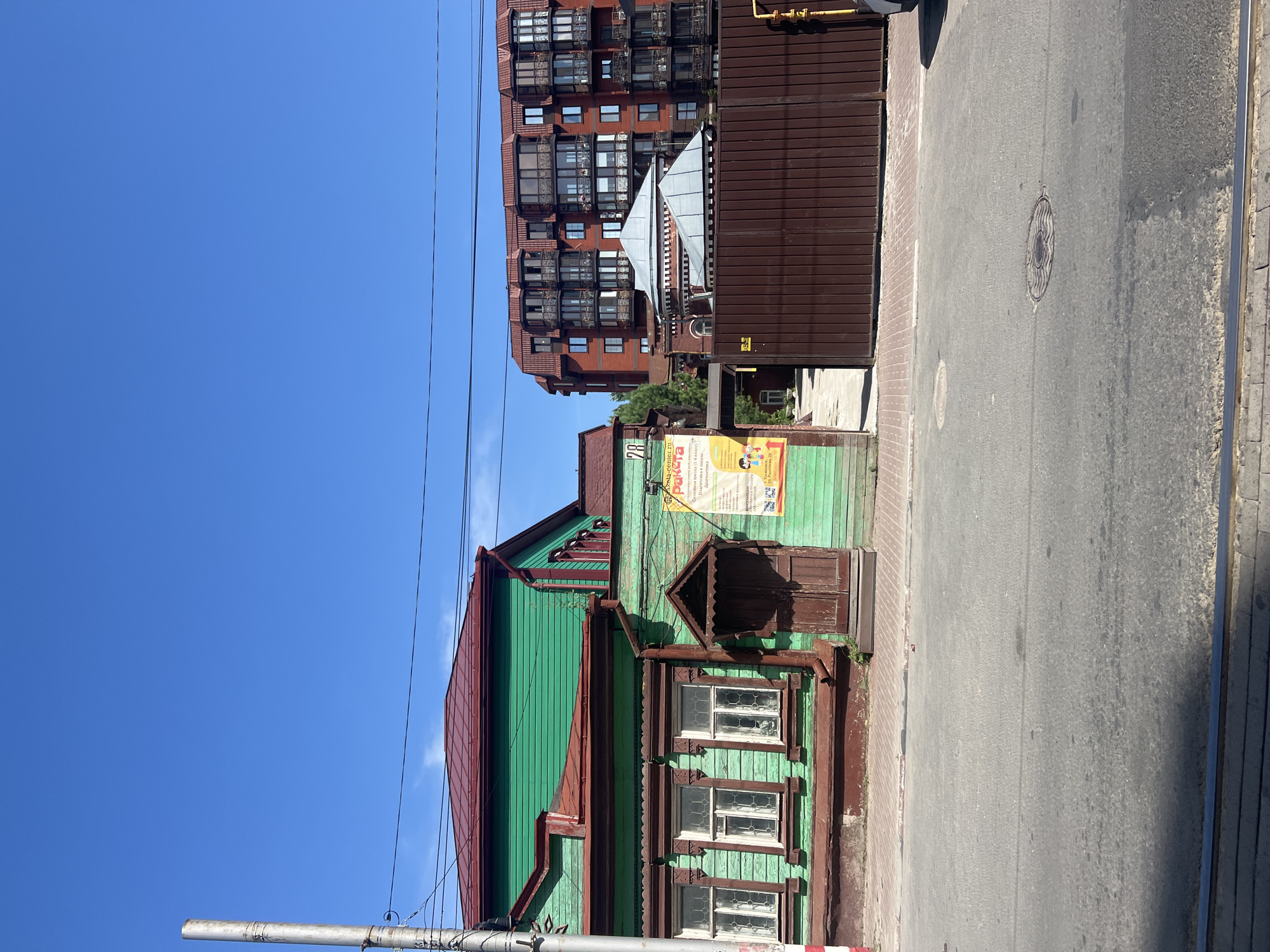 In Ulyanovsk, Russia, on a bright, sunny day, an image captures a street scene juxtaposing old and new architecture. On the left, a vibrant green wooden house with a red roof stands prominently. Its ornate wooden trim around the windows and a small, dark wooden porch with a decorative roof add character. A weathered wooden door is centered on the porch. Beside the house, a yellow poster is attached to the wall.
To the right of the wooden house, a modern, multi-story brick apartment building dominates the background. Its facade is adorned with numerous balconies, many featuring glass enclosures. In front of the apartment building and extending towards the viewer is a tall, dark brown metal fence. Between the wooden house and the fence, a circular-shaped building with a grey metal roof is partially visible. The foreground shows a paved street with a visible tram track. A manhole cover is set into the asphalt. The sky is a clear, bright blue.