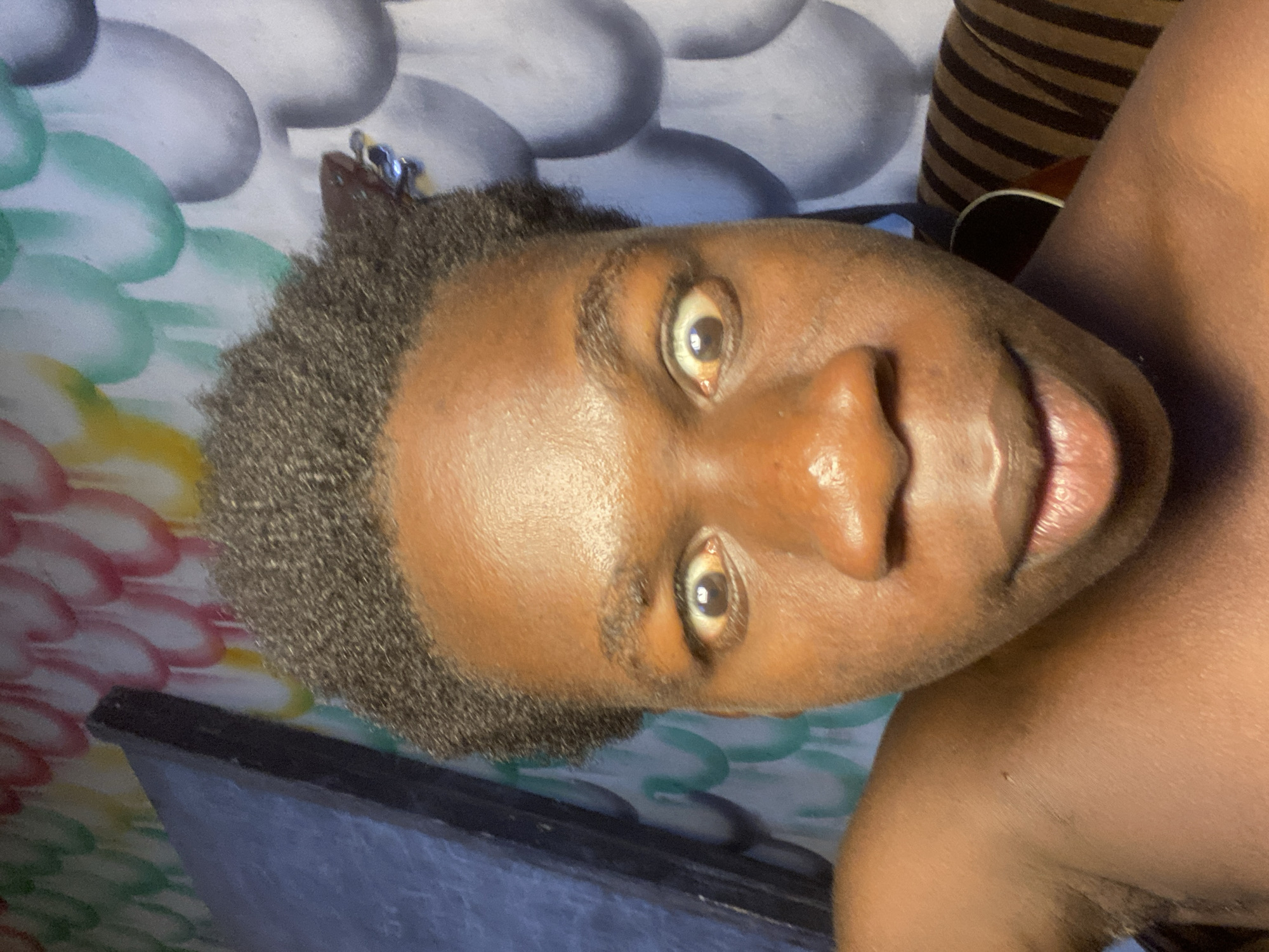 A close-up, rotated portrait of a dark-skinned individual with short, dark gray hair. The person's eyes are open and focused toward the camera. The background features a patterned surface in blue, green, white, and yellow hues. A dark object is visible behind the individual's right shoulder. The image's origin or presence in Mararaba, Nigeria, is not determinable from the visual information available.