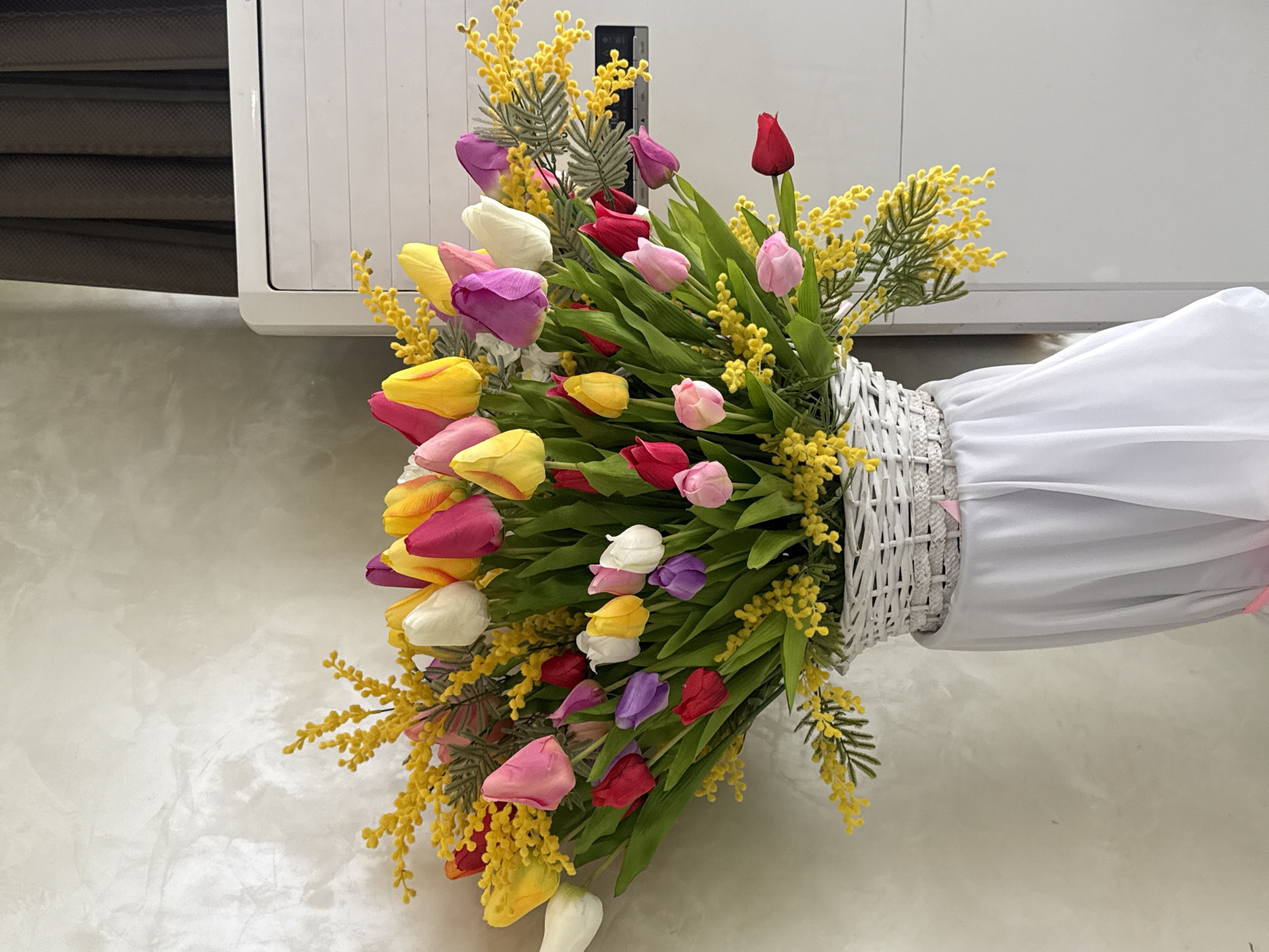 A high-angle, medium shot depicts a bouquet of colorful tulips and yellow mimosa flowers arranged in a white wicker basket. The bouquet is oriented horizontally, with the flowers extending to the left side of the frame and the basket wrapped in a white cloth on the right. The tulips display a range of colors, including pink, red, yellow, purple, and white. The mimosa flowers add a soft yellow touch, with their branch-like structure complementing the overall arrangement. The background consists of a white, horizontal surface and a light-colored wall with subtle textured patterns. The location appears to be indoors in the city of Unknown, Unknown. The lighting is bright and even, illuminating the bouquet and its surroundings. No people are visible in the shot. The focus is on the floral arrangement, creating a visually appealing composition.