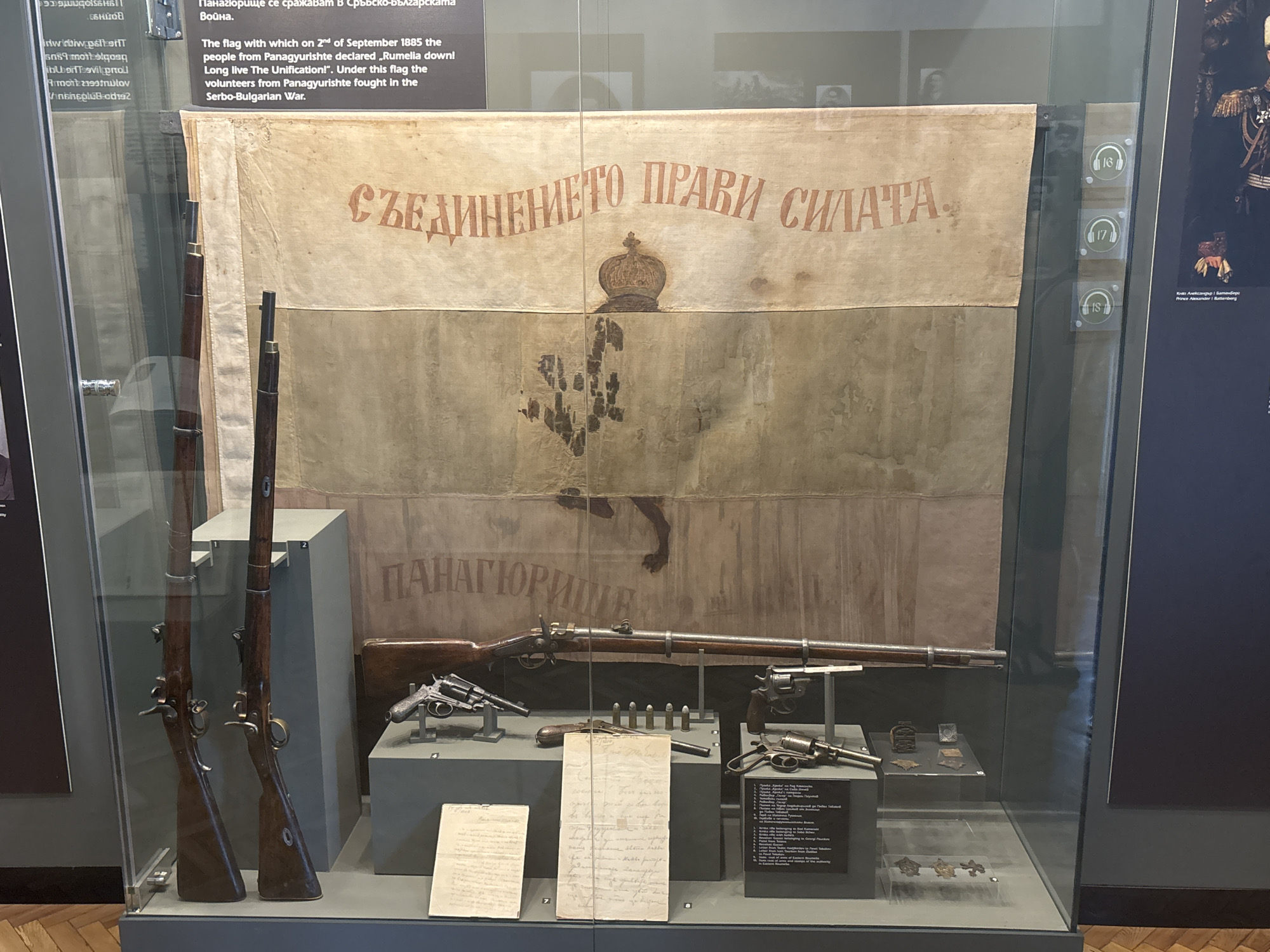 The image shows a museum exhibit featuring a flag and historical artifacts, likely from Panagyurishte, Bulgaria, though its specific location within the city is not visible. The central exhibit is a large, faded flag with Cyrillic text that reads "Съединението прави силата" (Unity makes strength). Below the text is an emblem, possibly a crown, and a darker, indistinct design on the lower portion of the flag.
To the left of the flag are two long rifles with wooden stocks and metal barrels. In front of the flag, on a display stand, are several firearms, including revolvers, and cartridges. Documents, appearing to be handwritten papers, are also displayed. To the right of the flag, there are small display cases with what appear to be medals or badges. The entire exhibit is enclosed in a glass display case, suggesting it is part of a museum. The background shows other exhibit elements and wall displays.
