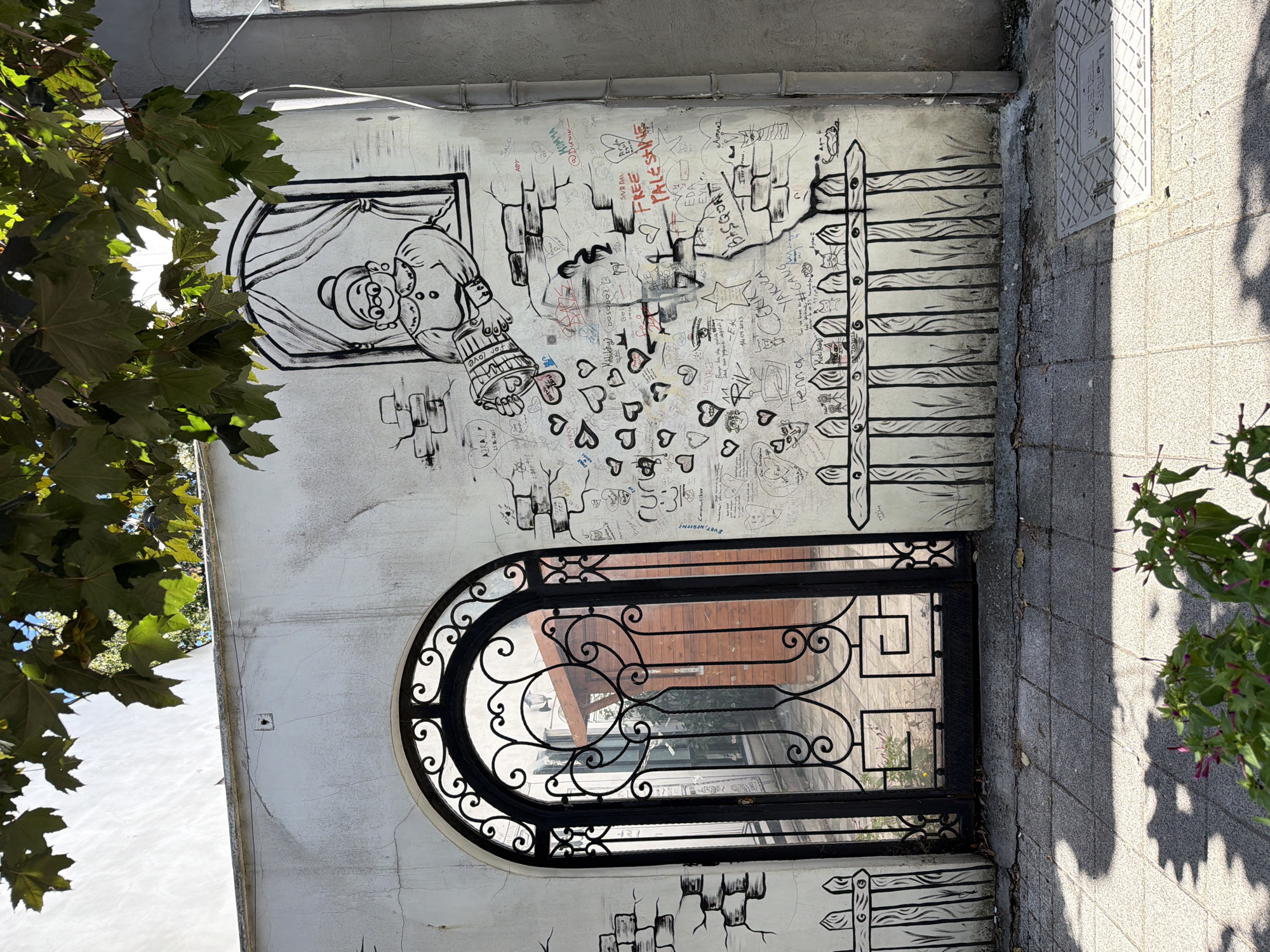 The image depicts a section of a white stucco wall in Kadıköy, Turkey, adorned with black and white graffiti and drawings. A tall, ornate black metal gate with an arched top occupies the left side of the frame, opening onto a courtyard with wooden structures visible beyond. The wall itself is decorated with a mural of an elderly woman peering out of a window, with hearts and a pail pouring love symbols falling from her hands. Various other drawings, including stick figures, cats, and political slogans like "FREE PALESTINE," are scribbled across the wall, some in red. The wall shows signs of age and some cracking. The ground in front of the wall is paved with light-colored square tiles, and shadows from overhead foliage fall across it. Green plants with small purple flowers are visible in the foreground. A metal drain cover with text is set into the pavement on the right side of the image. The overall scene suggests an urban environment with artistic expression and local culture.