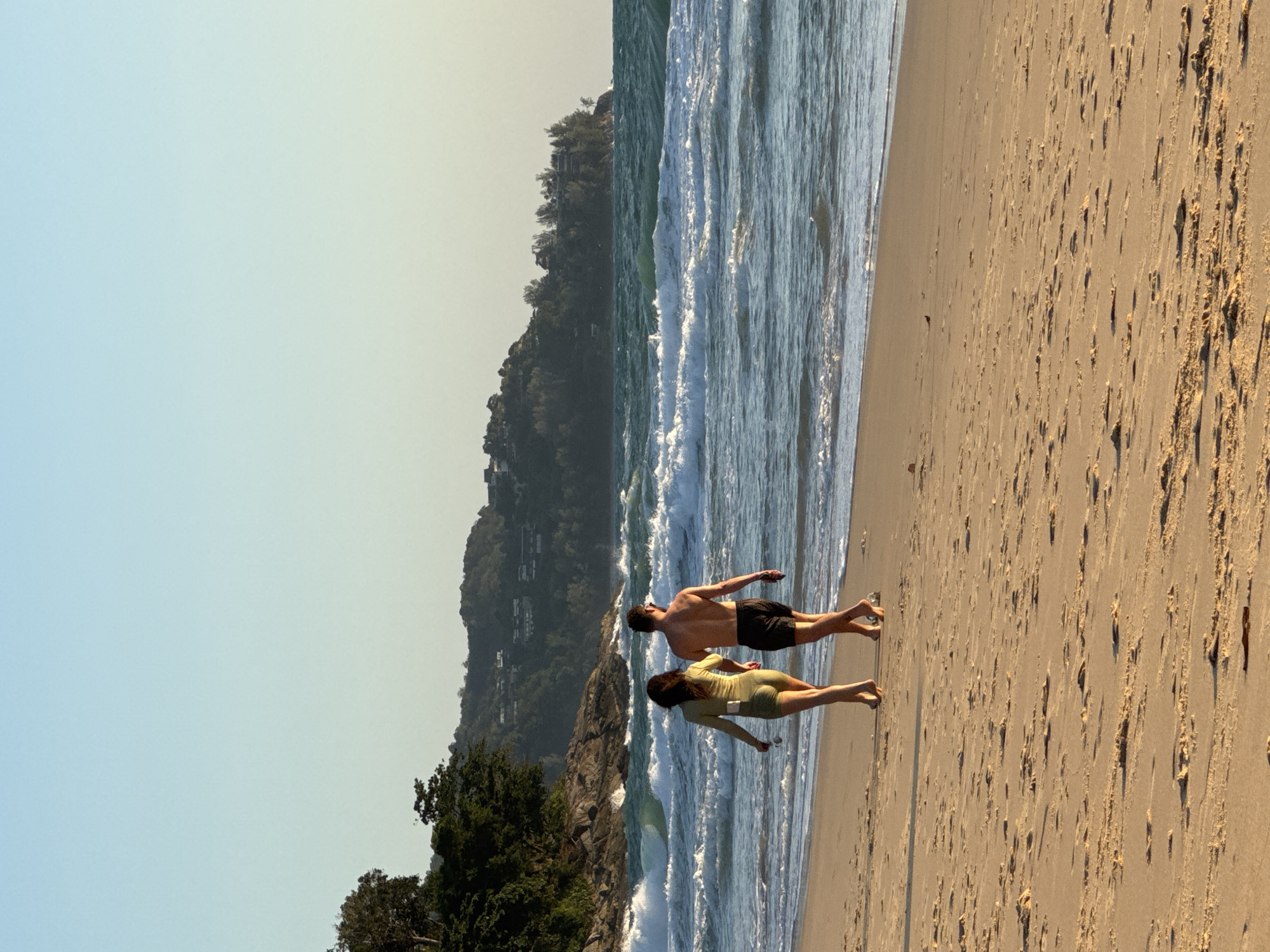 A man and a woman walk hand-in-hand along a sandy beach at sunset. The woman wears a pale green one-piece outfit, and the man wears black shorts. The ocean waves crash onto the shore behind them. In the distance, a tree-covered hill with buildings is visible under a clear, pale blue sky.