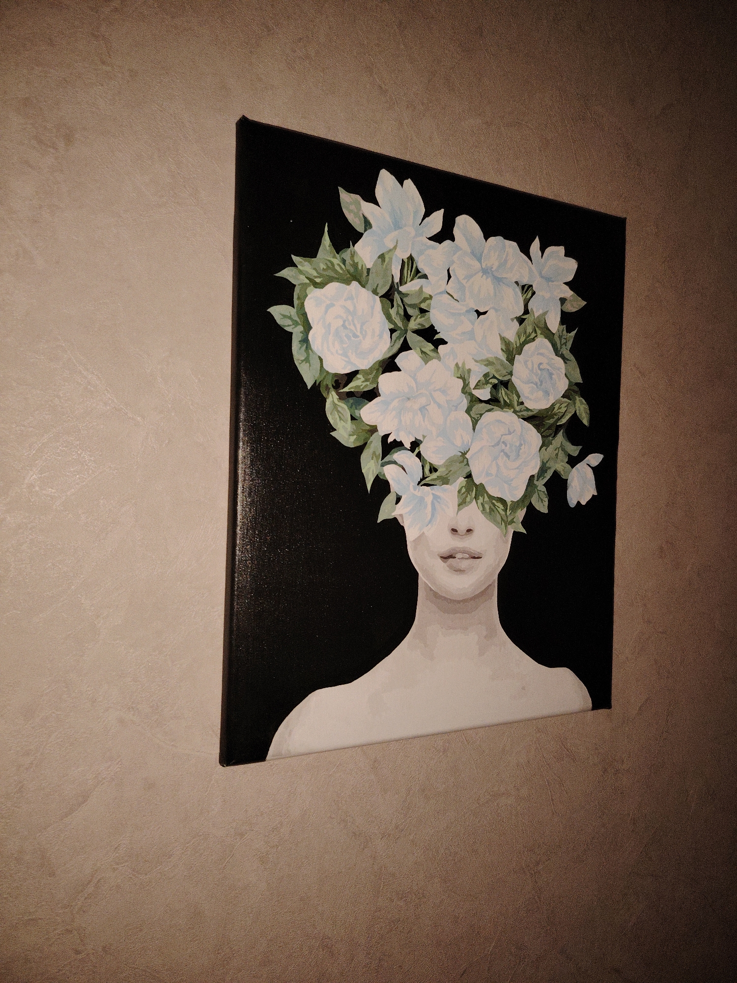The image shows a canvas painting hanging on a textured wall. The painting depicts a person with flowers covering their face and head. The person's skin is light-toned and the flowers are white and light blue with green leaves. The background of the painting is black. The wall is a beige-brown color with a slightly textured surface. The image is taken at an angle and the lighting is dim. The location is Марковский, Russia.