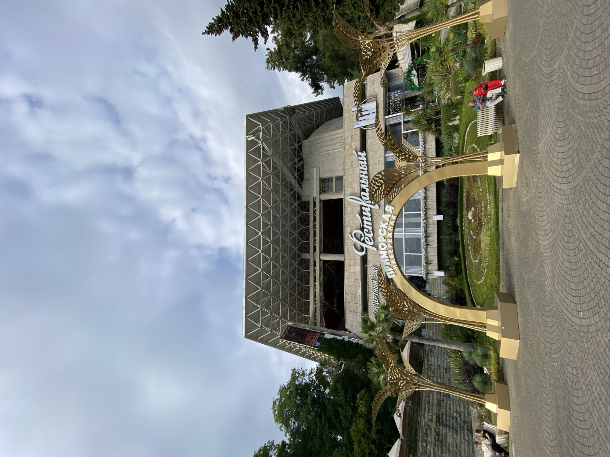 The image shows a large, modern building with a distinctive geometric roof structure, set against a cloudy sky. In the foreground, a large, ornate golden archway with stylized palm tree elements stands prominently. The archway has text on it. The scene appears to be a public space, possibly a park or promenade, paved with patterned stones.
There are a few people visible. Two individuals are seated on a bench near a circular garden area with manicured plants. Another person is partially visible in the lower right corner. The atmosphere seems relaxed, with the people on the bench appearing to be engaged in conversation or simply enjoying the surroundings.
The building in the background suggests a public venue, perhaps a concert hall or a cultural center, as indicated by the text on the archway which appears to be in Russian. Specifically, the text on the archway reads "ПРИМОРСКАЯ НАБЕРЕЖНАЯ" which translates to "Primorskaya Embankment" and "Фестивальный" which translates to "Festival". There is also text on the building itself, including what appears to be "Концертный зал" (Concert Hall).
The lighting suggests it could be daytime, but the overcast sky indicates potentially mild or diffused sunlight. The presence of palm trees and lush greenery suggests a warm climate, consistent with the given location context of Sochi, Russia. The overall impression is one of a well-maintained public area designed for leisure and cultural activities.