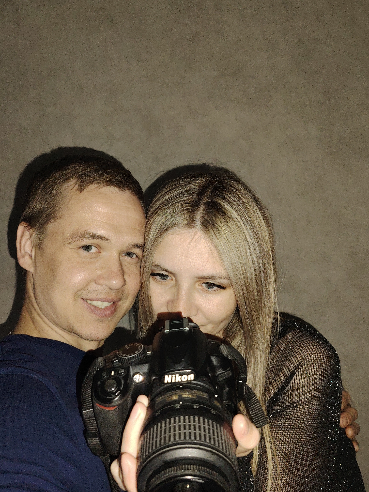 A portrait captures a man and a woman posing intimately together indoors in Chaikovsky, Russia. The man, positioned on the left, has short brown hair and blue eyes, and smiles warmly at the viewer, revealing his teeth. He is wearing a dark blue t-shirt. His left arm is wrapped around the woman beside him.
The woman, on the right, has long blonde hair with darker roots and blue eyes. She is focused downwards, looking at a black Nikon DSLR camera that she holds in the foreground. Her expression is one of concentration, possibly adjusting the camera or reviewing a shot. She wears a sheer black top with a subtle sparkling pattern. The brand name "Nikon" is clearly visible on the camera body.
The background consists of a plain, textured, light brown or grey wall, indicating an indoor setting. Prominent shadows are cast on the wall behind the couple, suggesting a direct light source. The image conveys a sense of closeness and happiness between the two individuals.