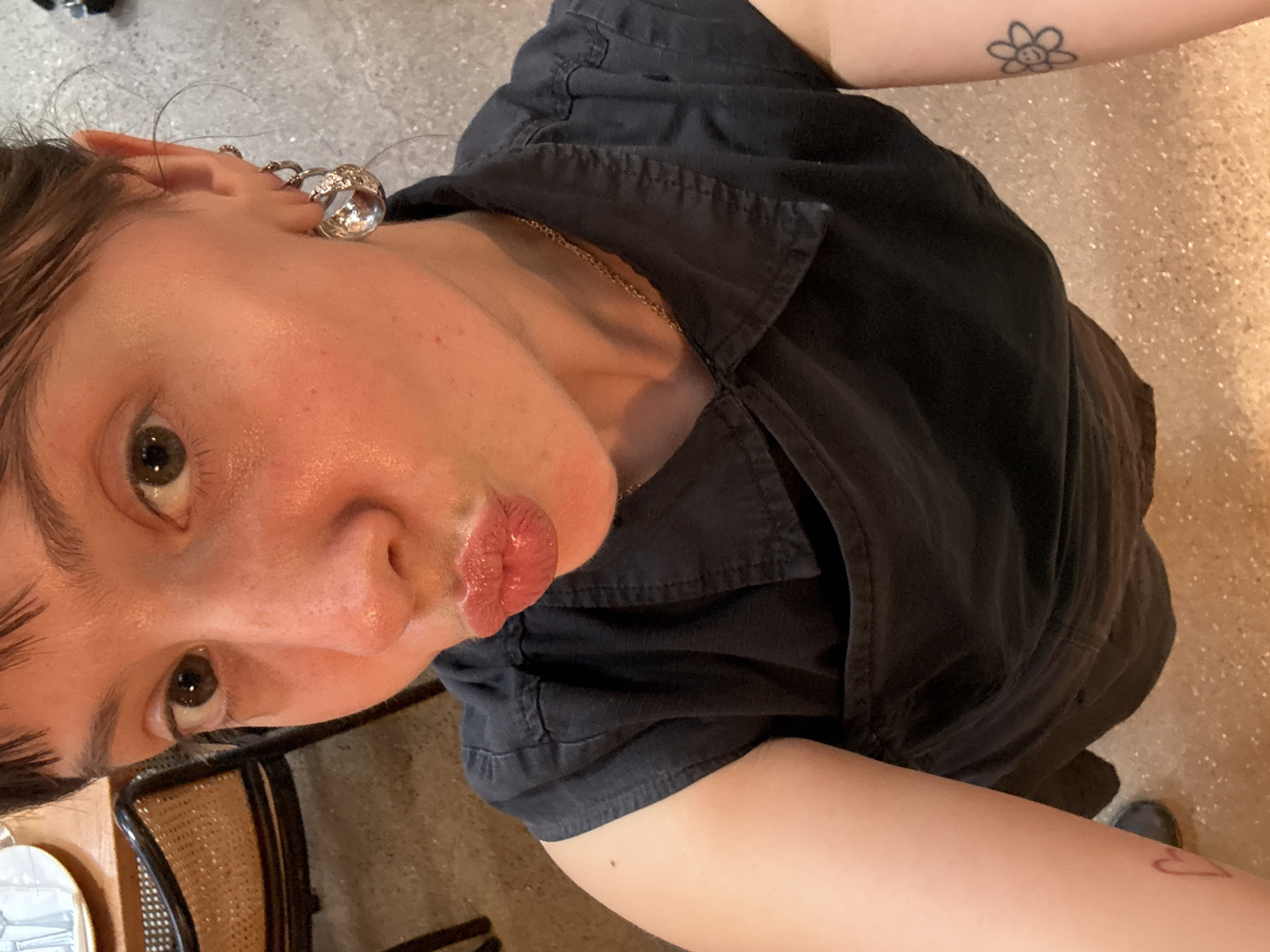 A young individual is captured in a high-angle selfie, looking downwards towards the camera with a playful expression. The subject, likely female, has fair skin, dark brown or hazel eyes, and short, dark hair. Their lips are puckered in a "kissy face," appearing glossy.
They are wearing a black, short-sleeved button-up shirt with a collar, and a thin gold chain necklace is visible around their neck. Notable accessories include large, shiny silver spherical hoop earrings on their left ear. Several visible tattoos include a black outline flower with a smiling face on their right upper arm and a red outline heart on their left forearm.
The setting appears to be indoors, possibly a cafe or restaurant. Parts of a light-colored, reflective floor with a sparkling texture are visible, along with a portion of a chair with a rattan-style weave and a table surface. The lighting suggests an indoor environment. The overall mood is casual and expressive. No explicit text is visible in the image. The location context provided is London, United Kingdom.