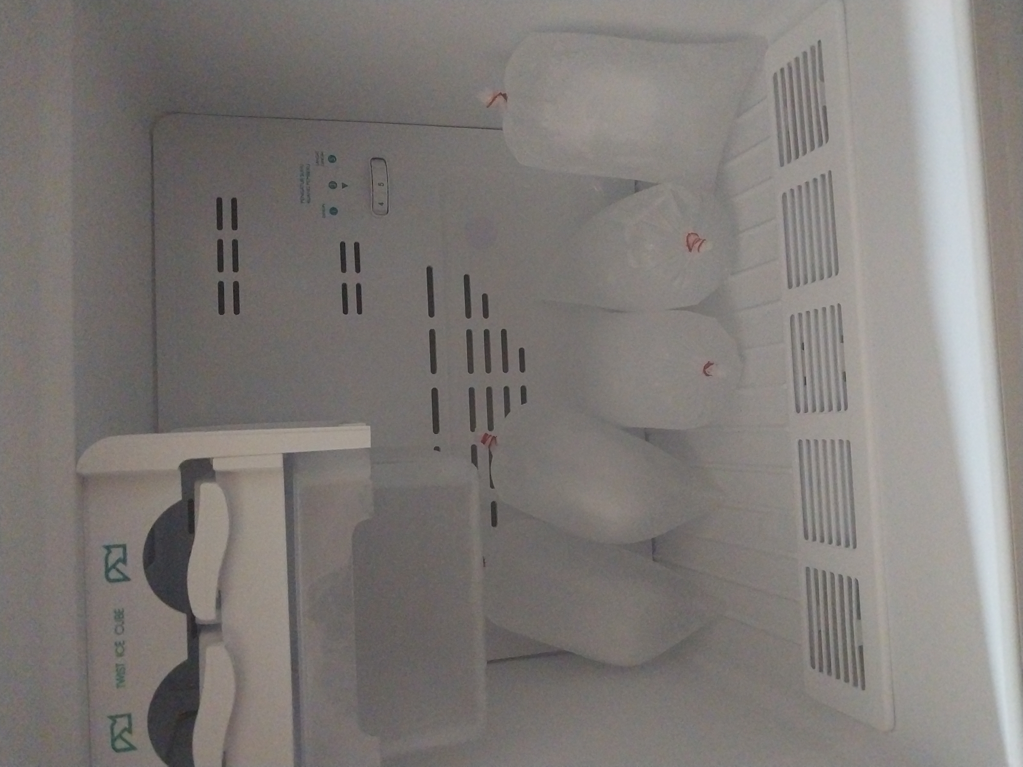 The image shows the inside of a refrigerator. In the top left is an ice cube maker with two dark gray levers and two white handles, along with a compartment labeled "TWIST ICE CUBE". Below this is a clear plastic bin. The main body of the refrigerator interior is visible, with several bags of ice on the bottom shelf. The bags are clear plastic with red ties. There are horizontal vents on the back wall and on the bottom shelf. A control panel with buttons and a dial is visible on the back wall. The scene appears to be a typical household appliance. There is no specific indication of the location being Air Tiris, Indonesia; this information cannot be determined from the image alone.
