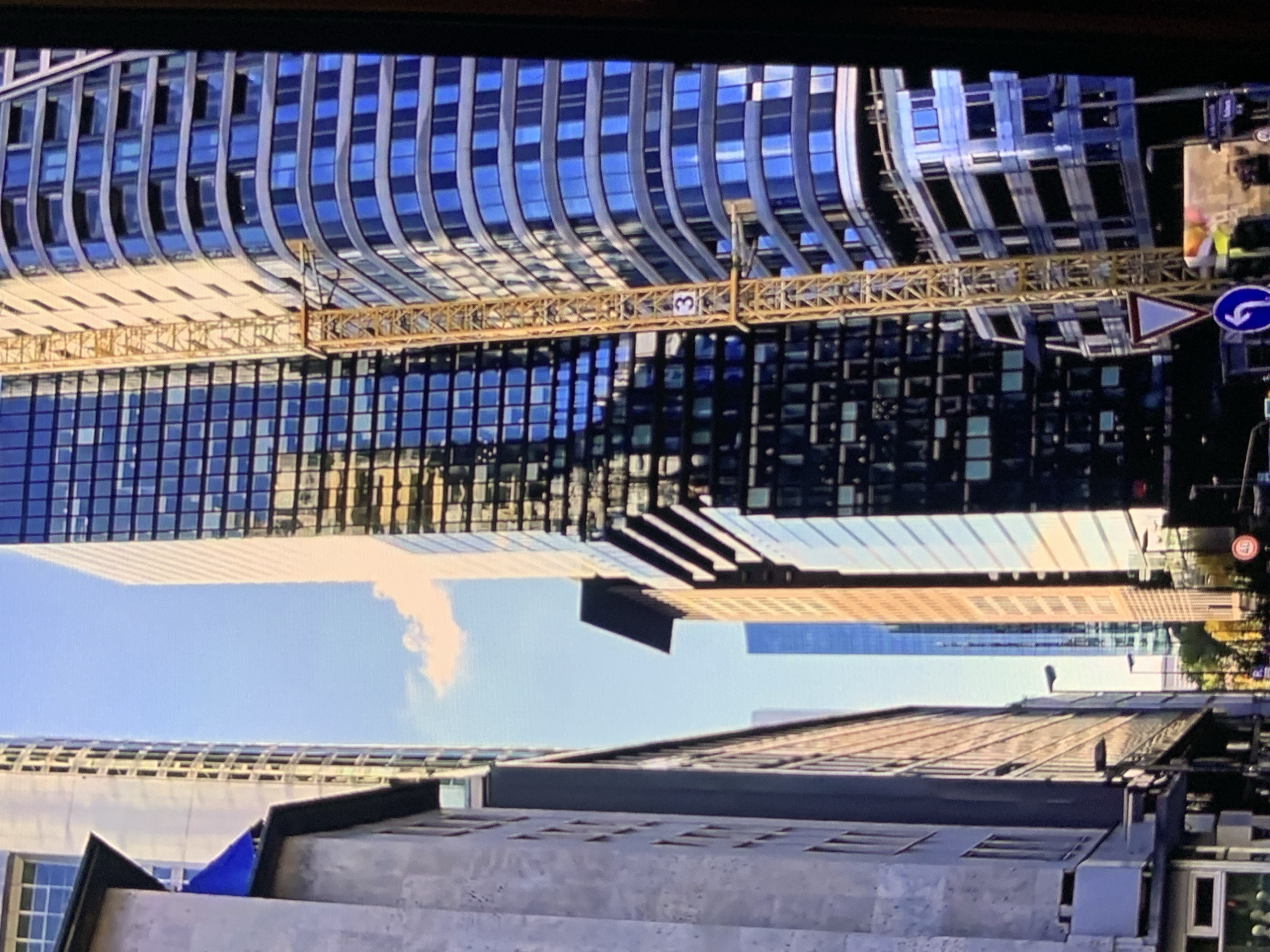 The media file shows an urban scene dominated by modern, tall buildings with blue glass facades. A yellow construction crane is prominently positioned across the middle of the frame, spanning between two buildings. The crane has the number "3" visible on one of its components. The sky above is bright blue with a few wispy clouds, suggesting it is daytime. Reflections of the buildings and the sky are visible on the glass surfaces.
The perspective is from a low angle, looking up at the towering structures and the crane. The buildings exhibit a curved architectural style. In the foreground, another building with a concrete-like texture and a blue tarp or flag is visible, partially obscuring the view of the sky. The scene suggests ongoing construction or development in a city. There are no visible people.
The location context provided is Stavropol, Russia. Time of day appears to be midday due to the bright sunlight and clear sky. Weather is fair. Visible text includes the number "3" on the crane and what appears to be a street sign with a blue circular arrow and a triangular warning sign. The overall mood is one of urban development and progress.