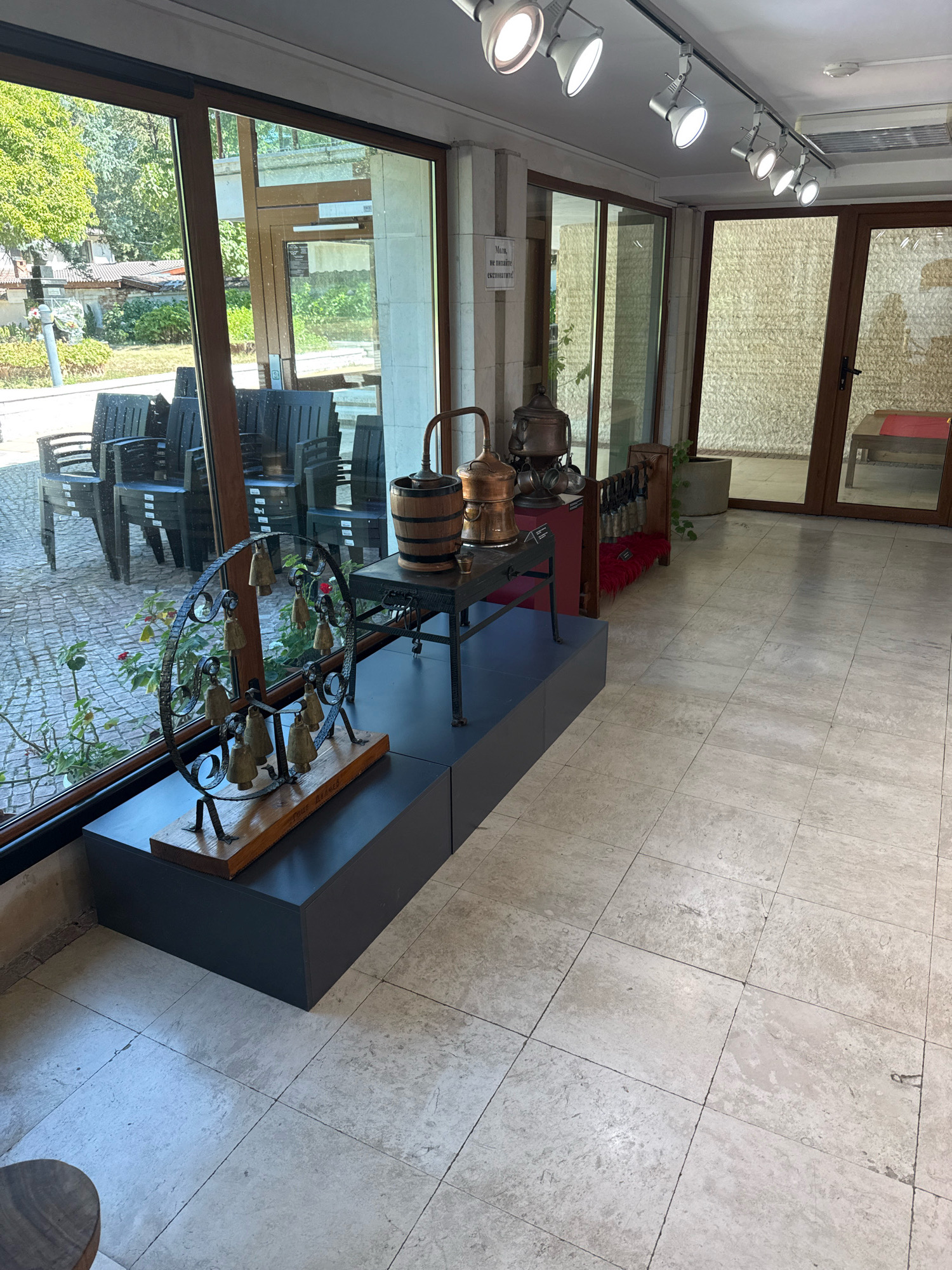 The image depicts the interior of what appears to be a museum or exhibition space in Pravets, Bulgaria, likely connected to the life and work of Todor Zhivkov. The foreground is dominated by a display featuring a circular metal structure with numerous small, brass-colored bells attached, mounted on a wooden base. This exhibit rests on a dark gray, rectangular pedestal. To the right of this display, a tall, dark metal table with wheels holds several antique copper distilling apparatuses. Behind these objects, through large windows, an outdoor scene is visible, showing paved ground and an arrangement of dark, modern-looking chairs, suggesting an exterior seating area. The interior walls are a light, textured stone, and the floor is tiled with large, light-colored travertine tiles. Overhead, a track lighting system illuminates the space. In the background, large glass doors lead further into the building, revealing more textured walls and potentially other exhibits. A plant is visible in a cylindrical pot to the right of the distilling apparatuses.